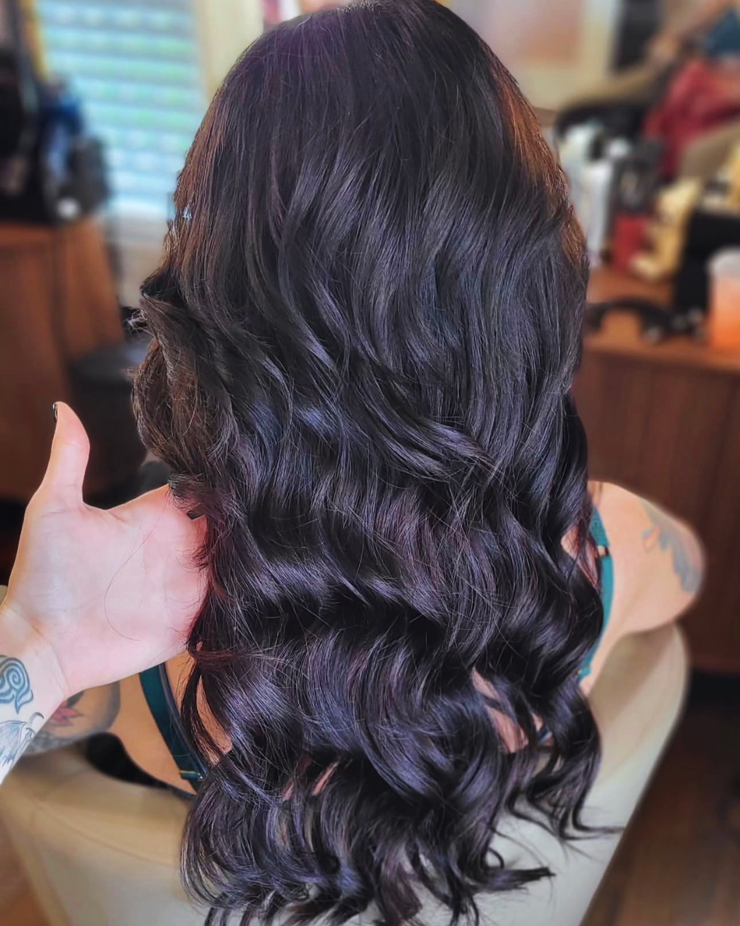A person with long, wavy dark purple hair sitting indoors, with a hand touching their hair.