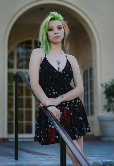 Young woman with vibrant green and pink hair standing outside a building with large windows and an arched entrance. She wears a black dress with a floral pattern and holds a railing.