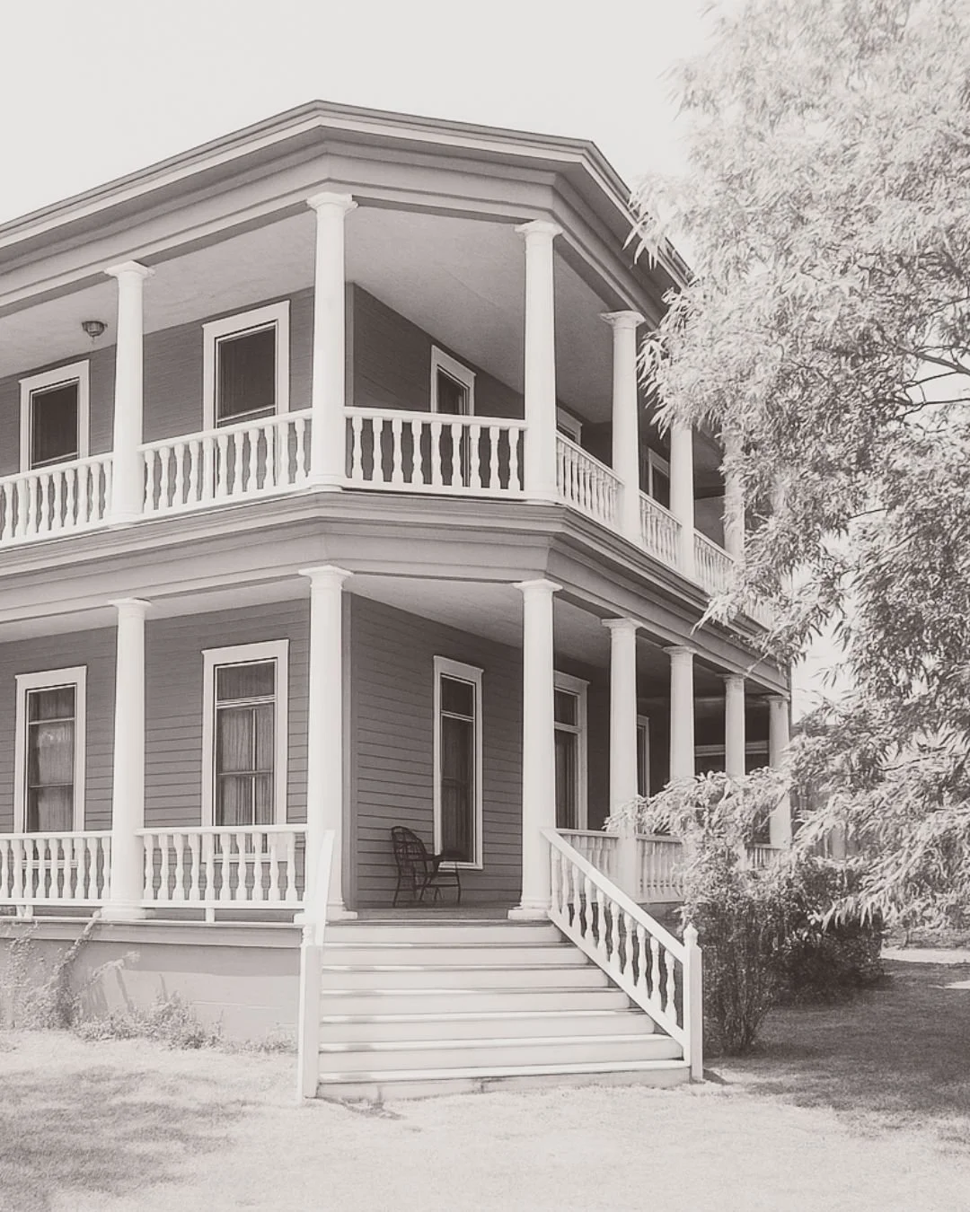 Black and white photo of a two-story house with a wraparound porch, columns, stairs leading up to the front door, and a bench on the porch. Trees partially visible on the right side.