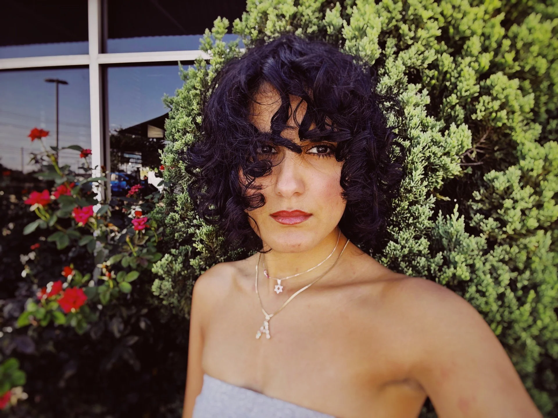 A woman with curly black hair and light makeup taking a selfie outdoors, standing in front of green bushes and a building with reflective windows.