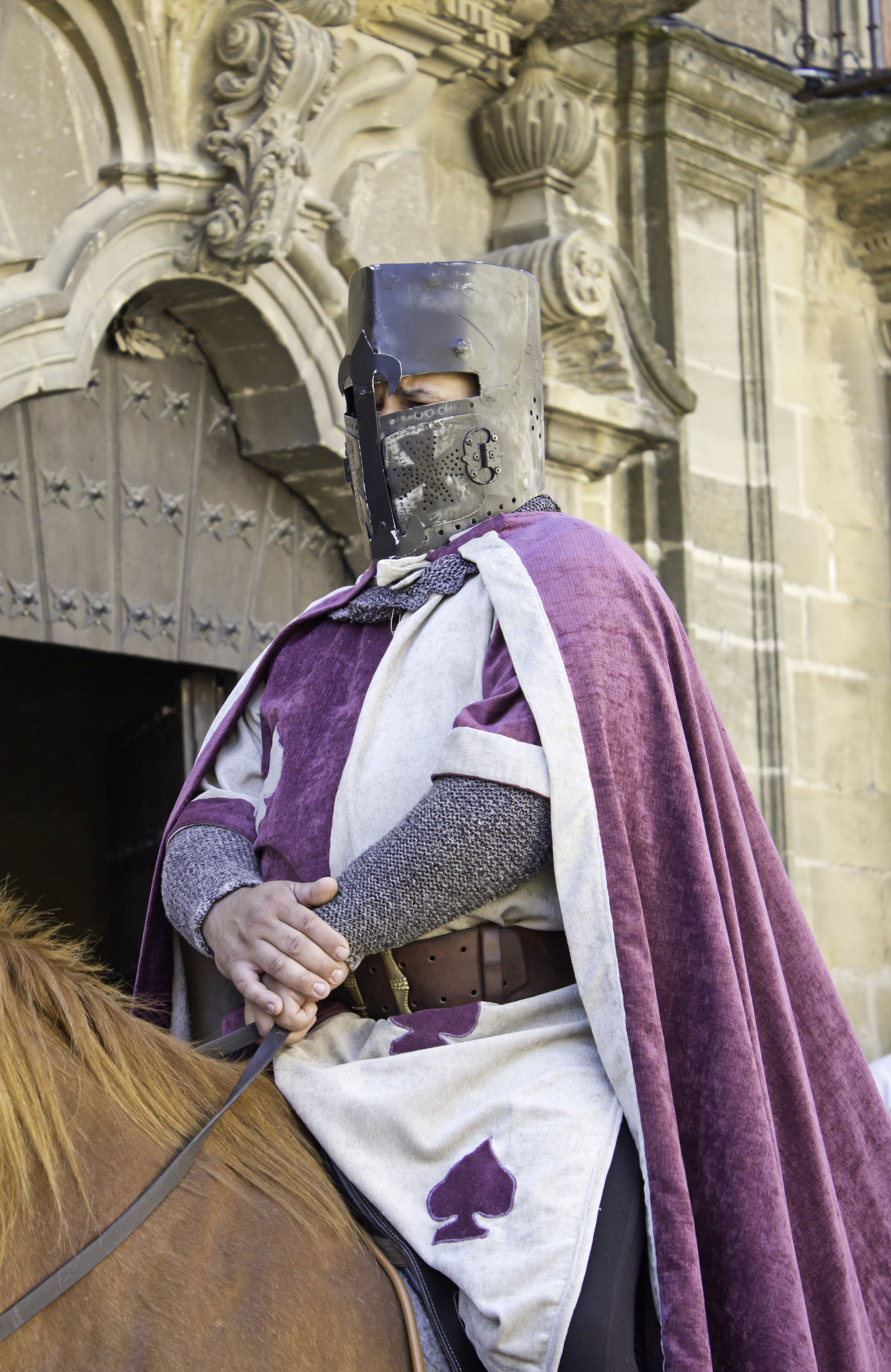 A person dressed in medieval knight armor with a helmet, riding a horse, and wearing a purple and white cloak with spade symbols, in front of a historic stone building.