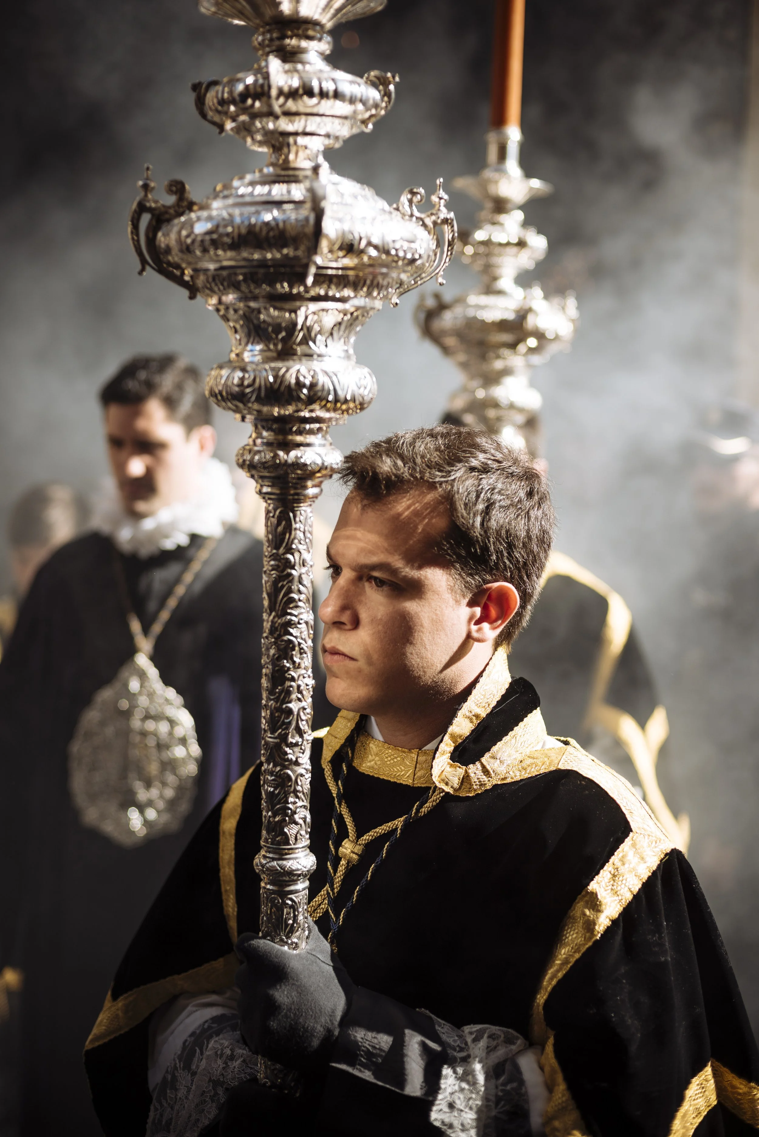 Young man dressed as a bishop holding an ornate silver processional cross, with other men in similar costumes in the background.