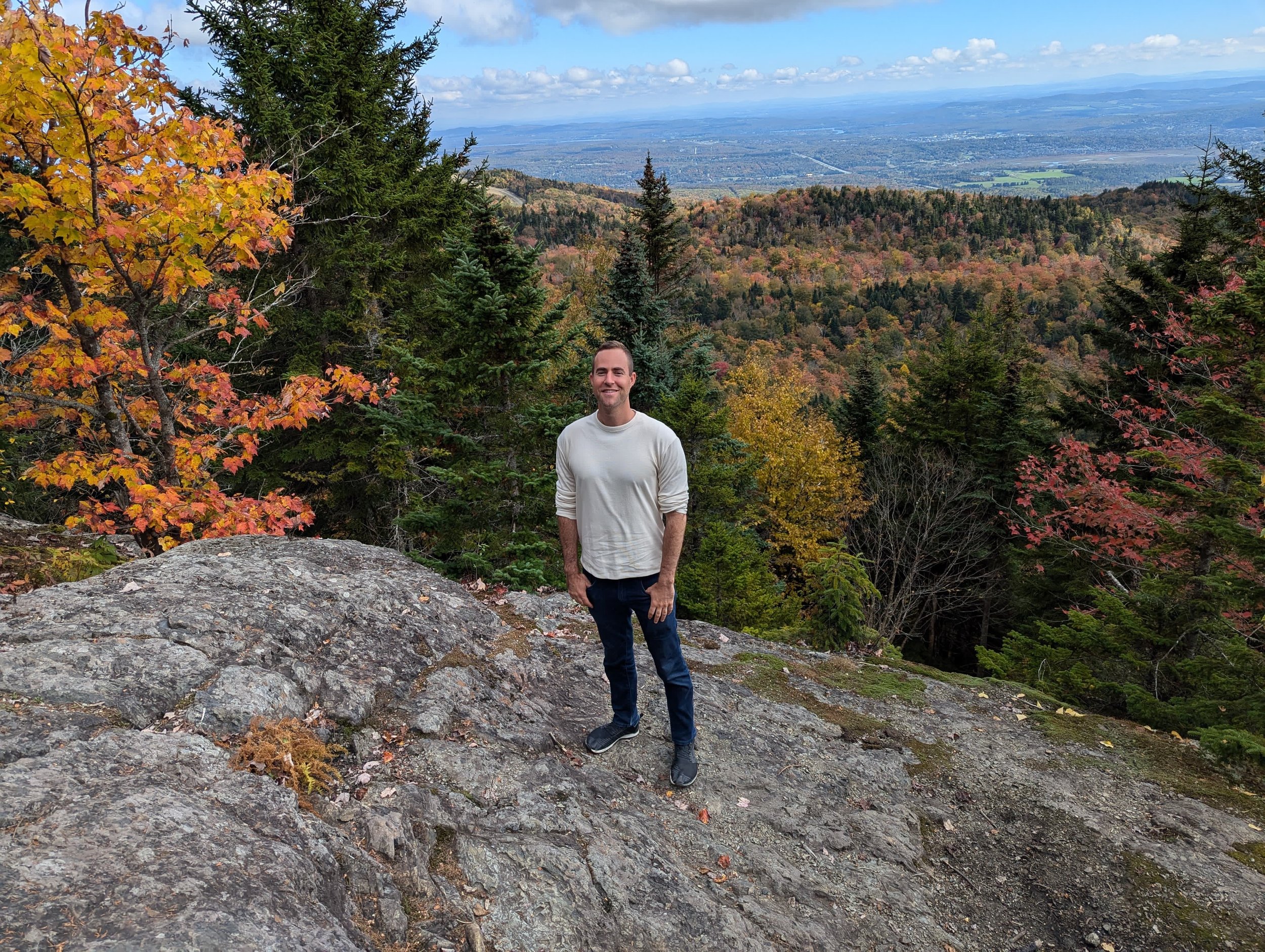 A man standing on rocky terrain among autumn trees with colorful leaves, overlooking a valley and distant mountains under a partly cloudy sky.