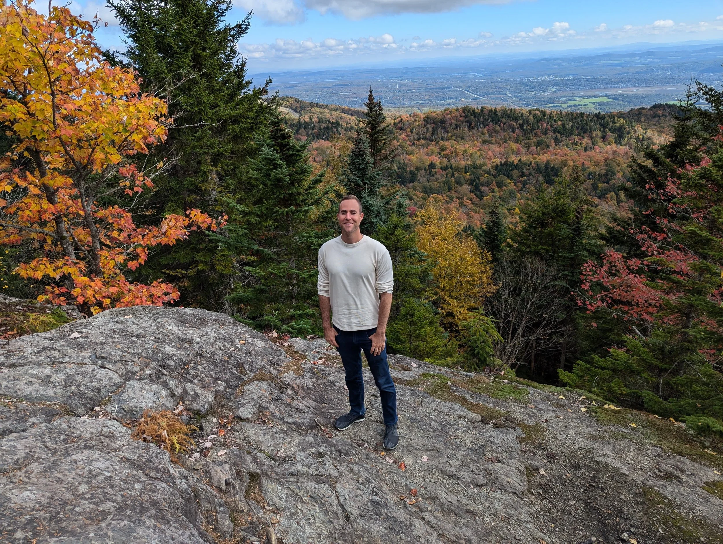 A man standing on a rocky outcrop surrounded by colorful autumn trees with a view of a valley and distant mountains in the background.
