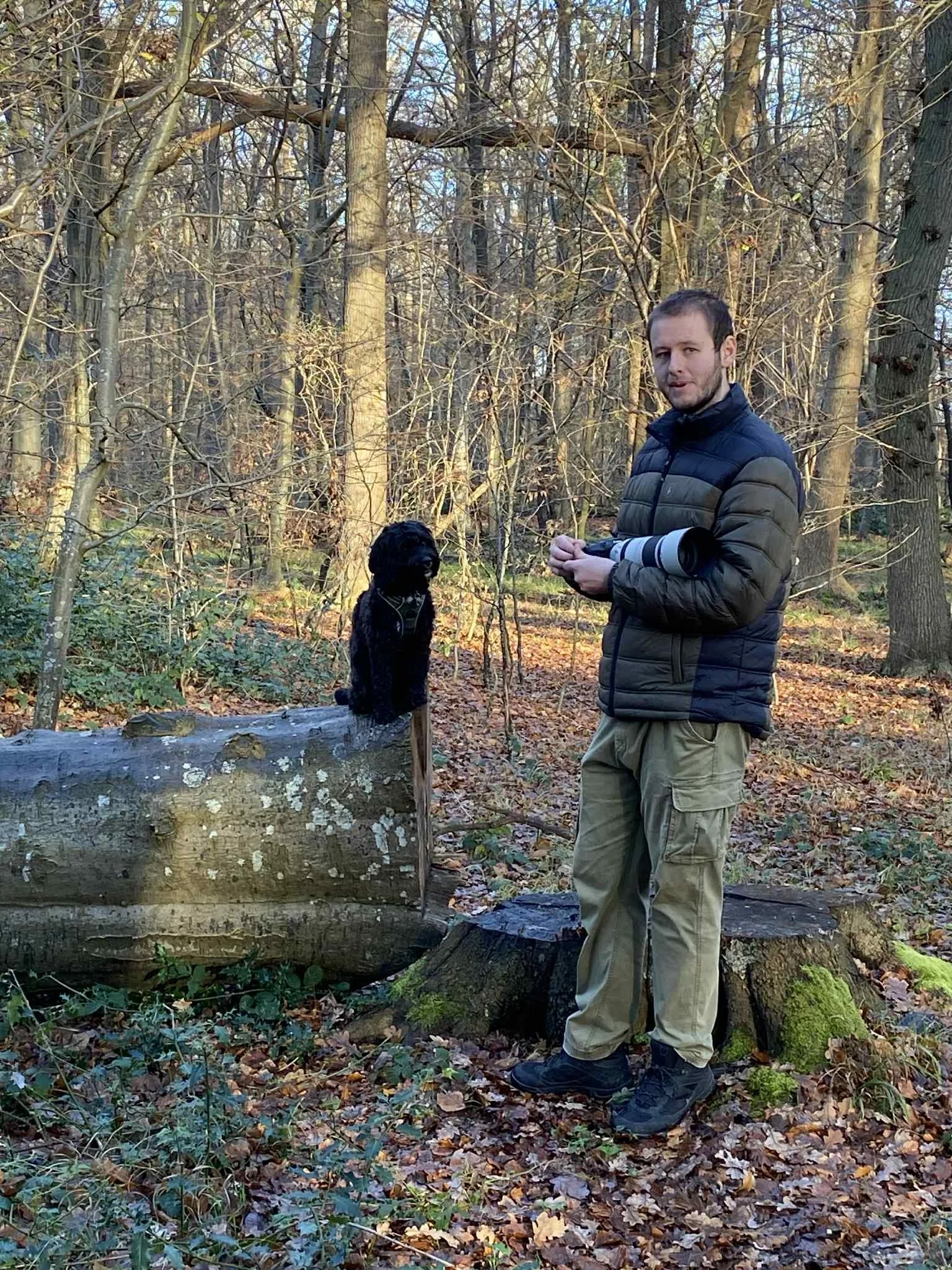 En mand står i en skov, holder et kamera med teleobjektiv, og der er en sort hund siddende på en stor træstub ved siden af ham.