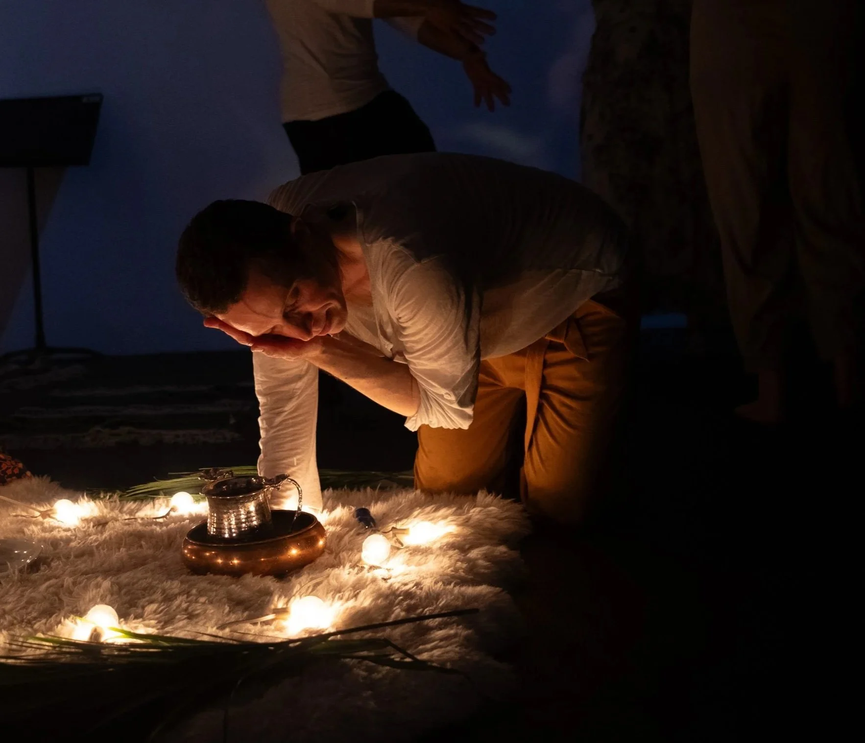 A person is kneeling on a fluffy white surface, cuddling a dog and looking at string lights and a jug or pitcher, with an outdoor backdrop of a blue sky and clouds during twilight.