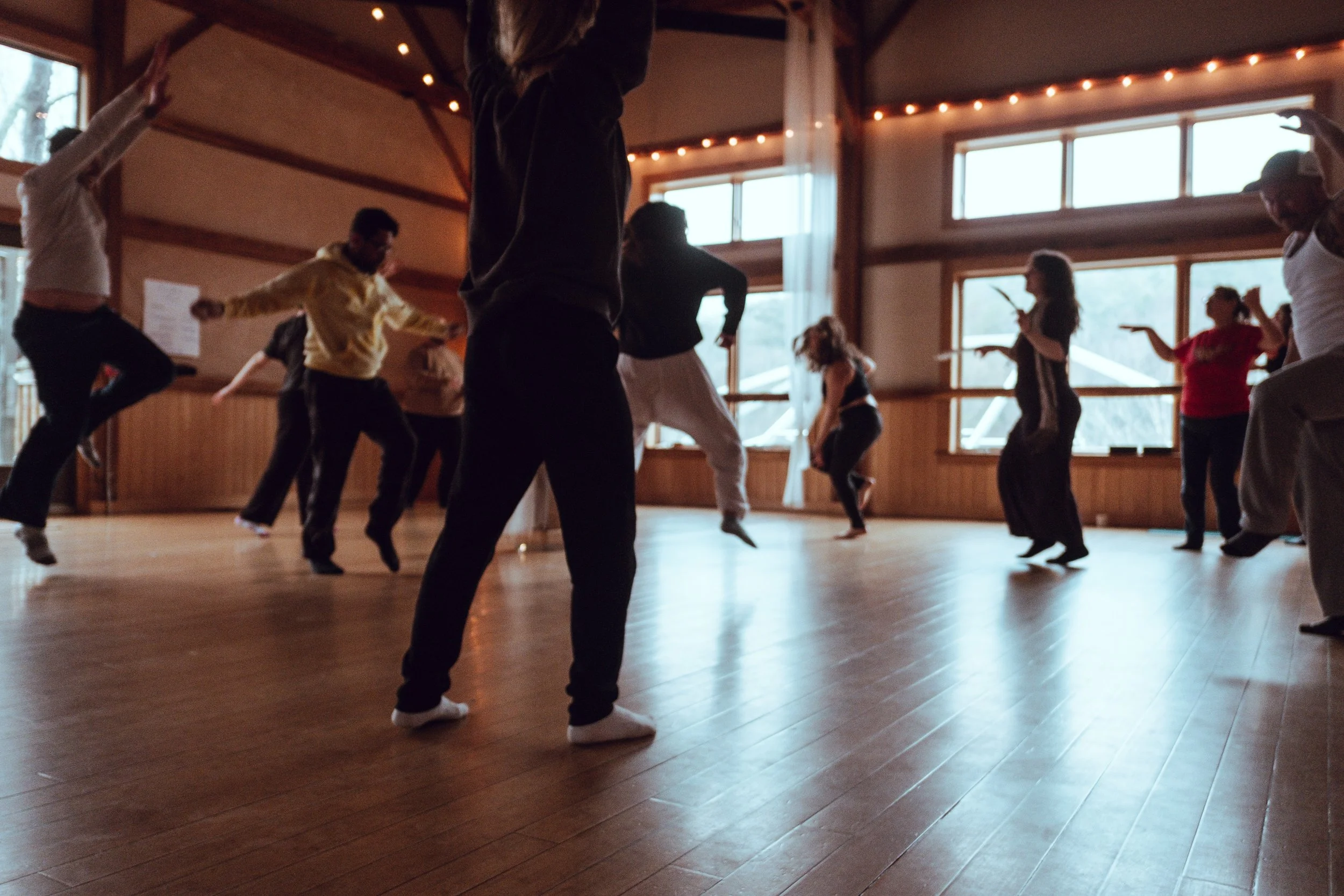 Group of people dancing together in a spacious wooden-floored room with large windows and string lights overhead.