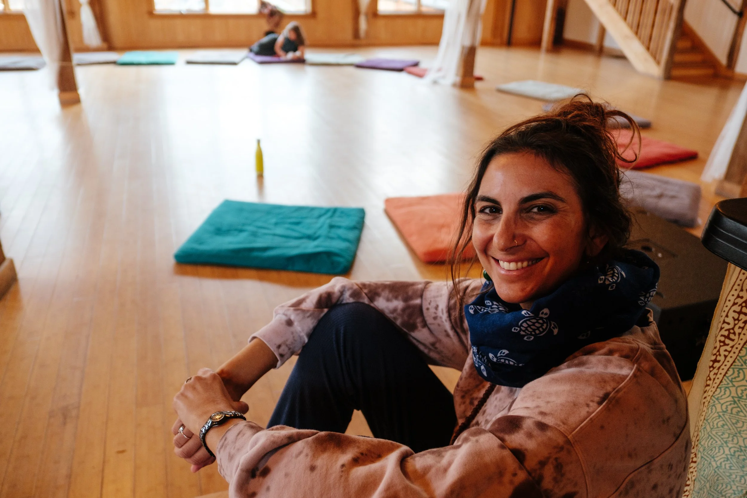A woman with dark hair smiling at the camera, sitting in a yoga studio with wooden floors, with colorful mats and two children practicing yoga in the background.