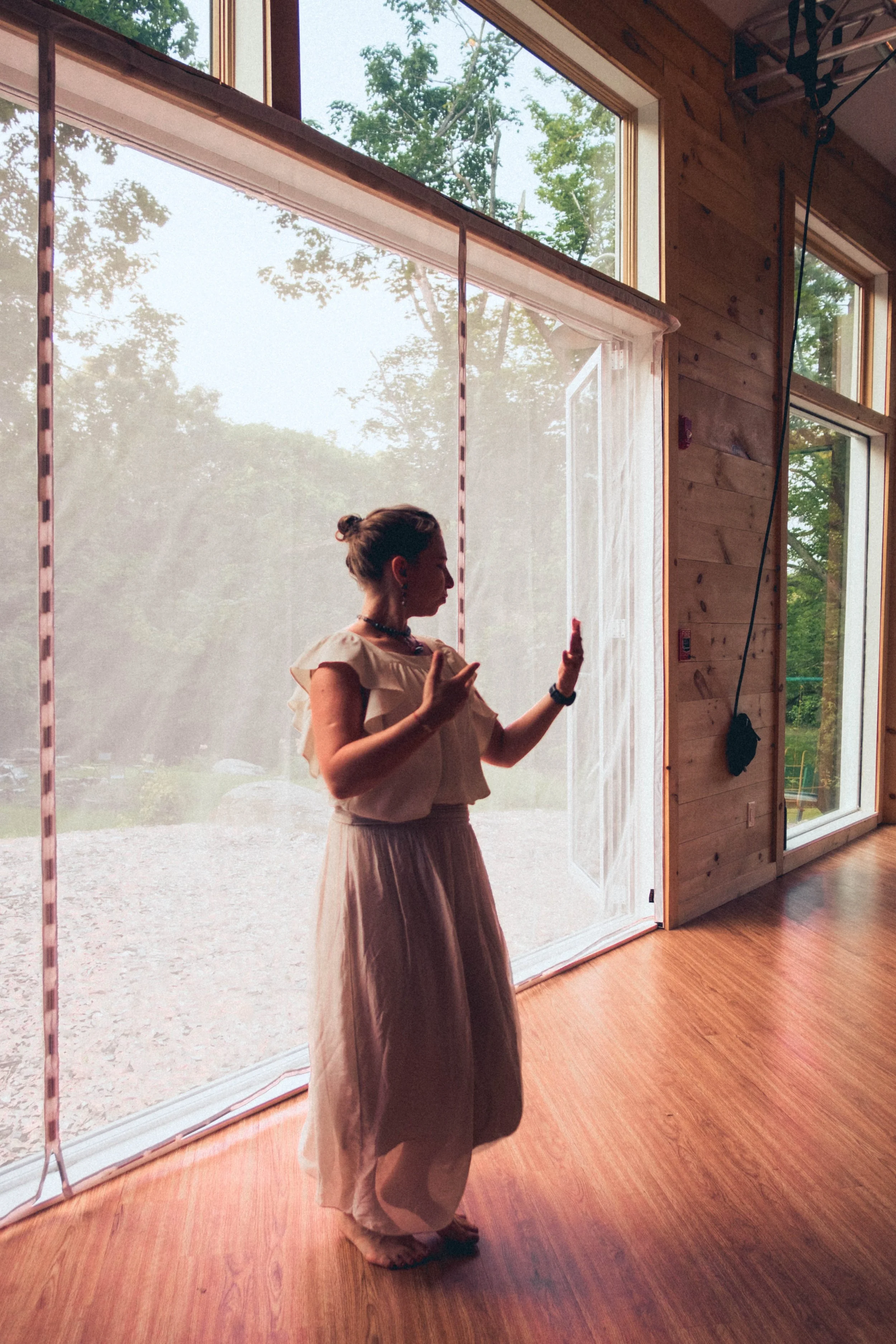 A woman standing barefoot indoors near large windows, gesturing with her hands, with trees and sky visible outside.