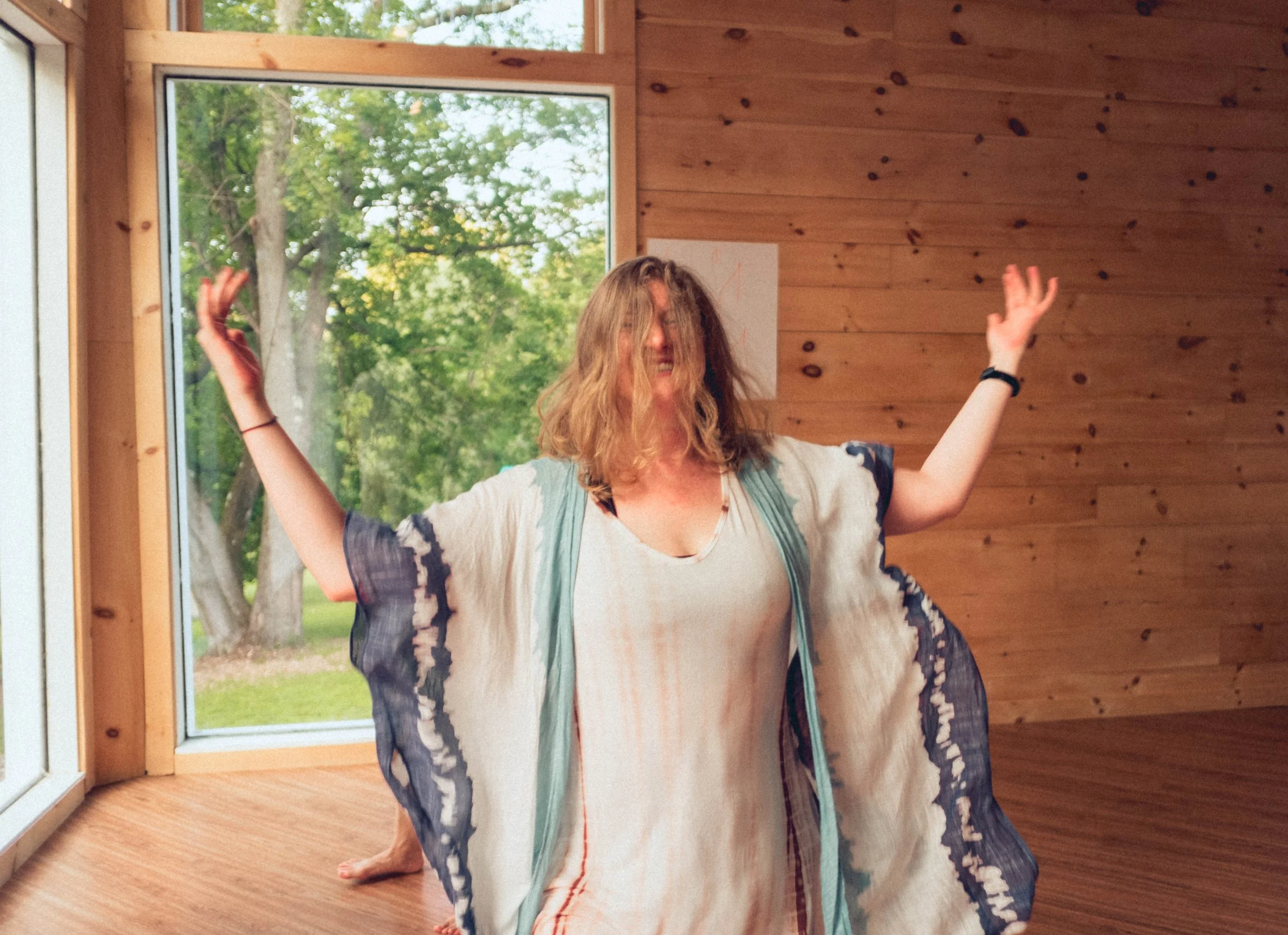 A woman with shoulder-length hair, wearing a flowing, colorful top, has her arms raised and appears to be smiling or laughing indoors next to a large window with a green outdoor scene.