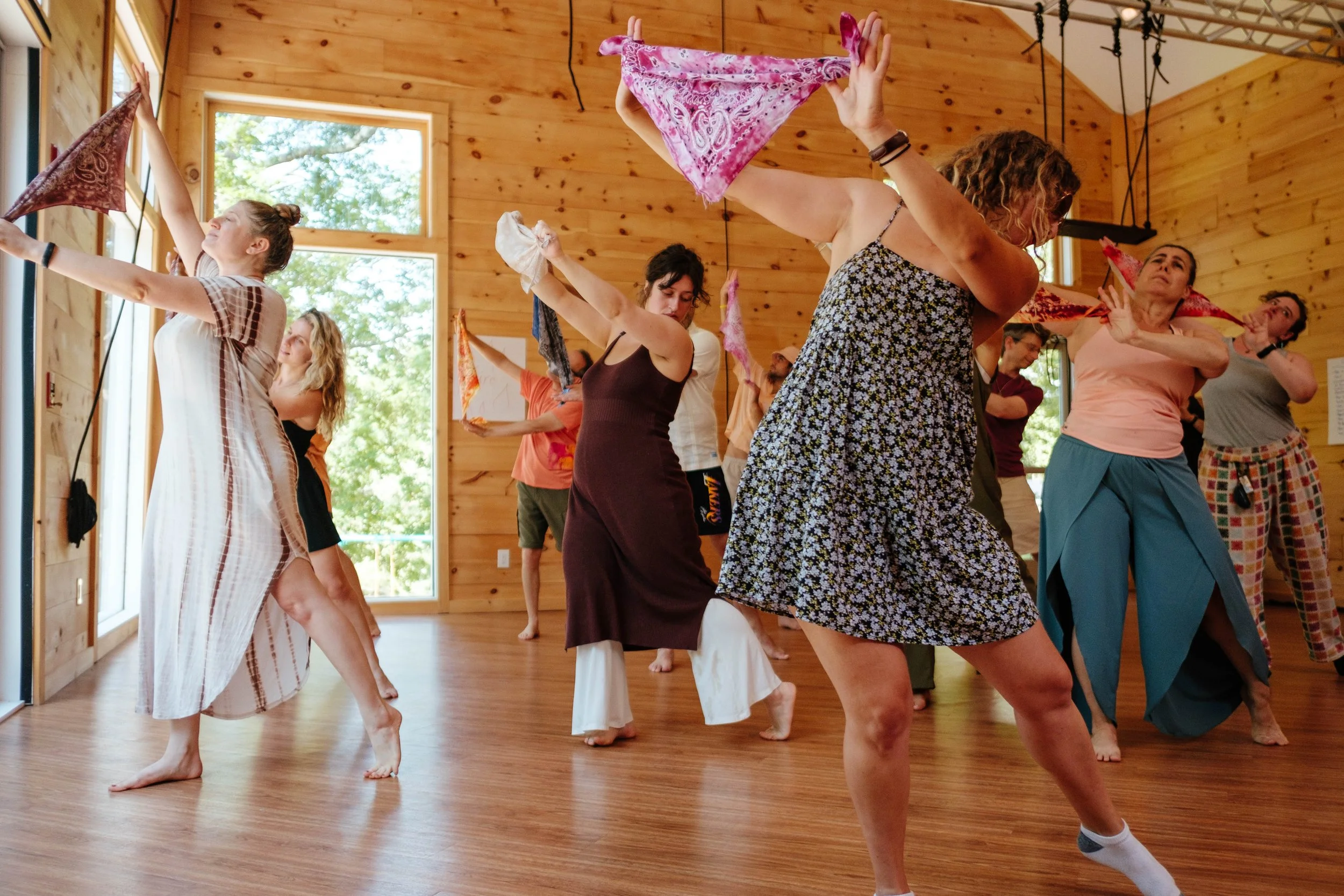 Group of women practicing dance or movement exercises in a wooden studio with large windows, some holding colorful scarves or fabric.