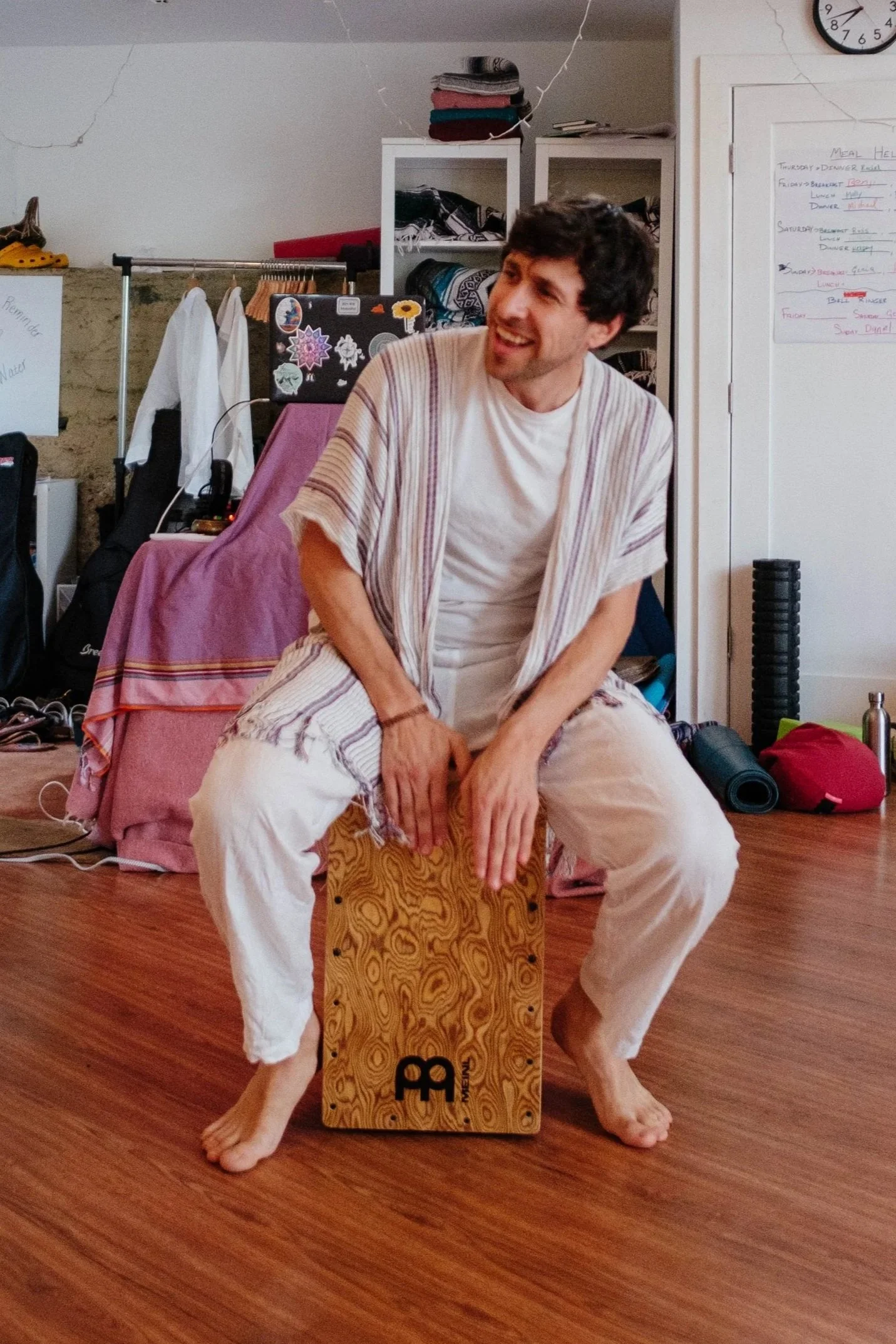 A man sitting on a wooden cajón drum in a room with various objects and whiteboards in the background, smiling and dressed casually.