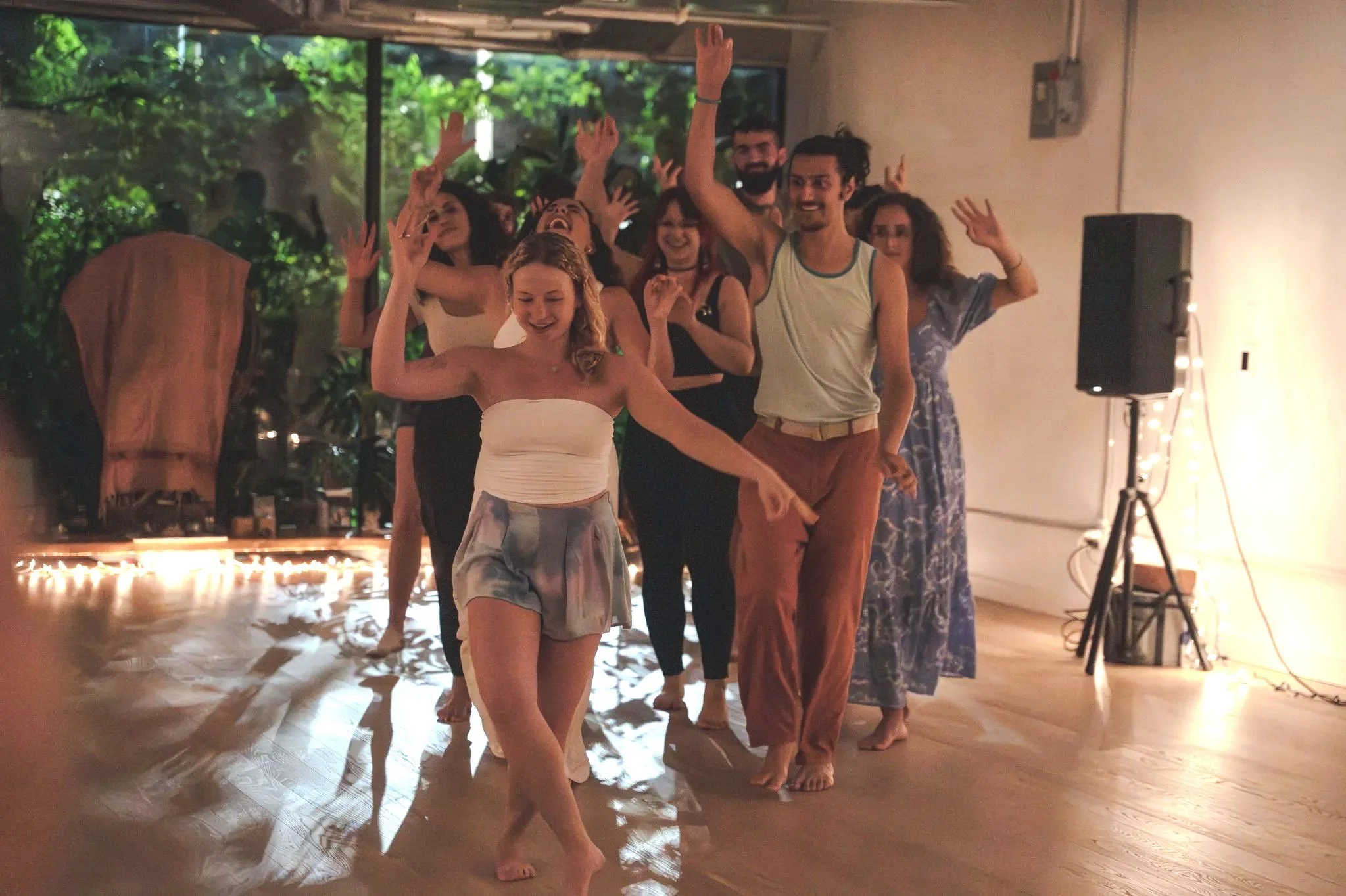 Group of people dancing and smiling at an indoor party with string lights and a large window in the background.