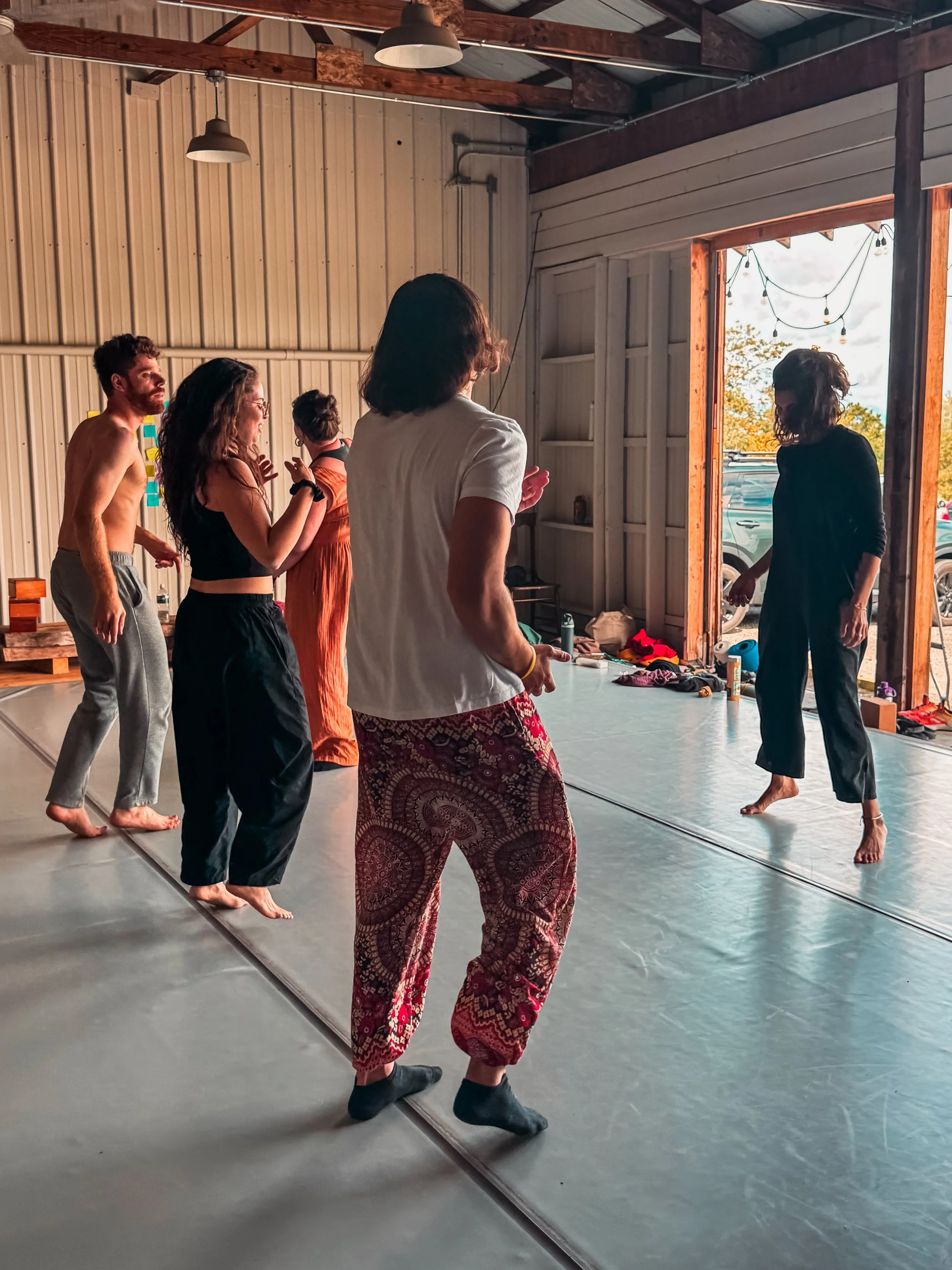 Group of people practicing yoga or dance in a rustic indoor studio, with a large open doorway showing an outdoor view with cars and trees.