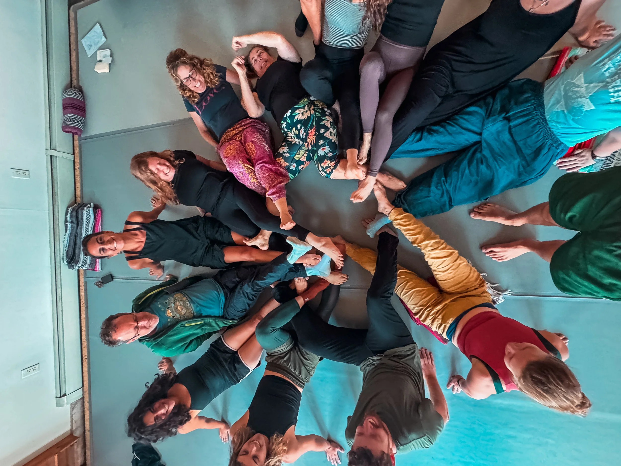 Group of people practicing acroyoga indoors on a blue mat, forming a human pyramid with some supporting others and some balancing.