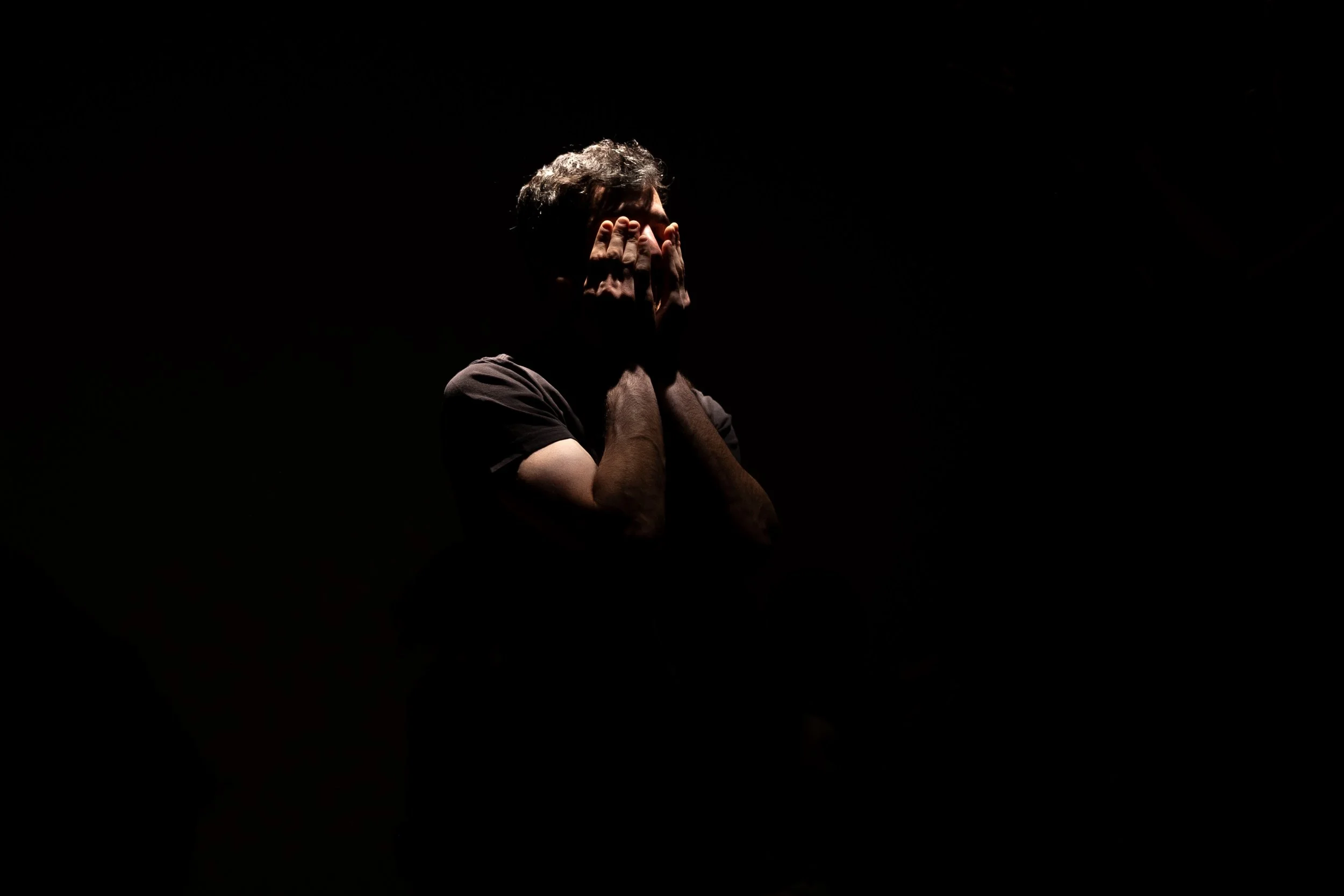 Person covering their face with hands in front of a dark background, low lighting.