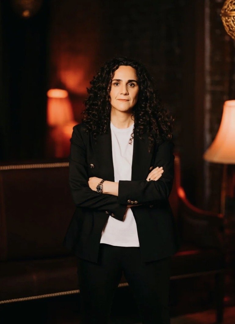 A woman with curly dark hair, wearing a black blazer over a white shirt, standing with crossed arms in a dimly lit room with warm lighting and lamps.