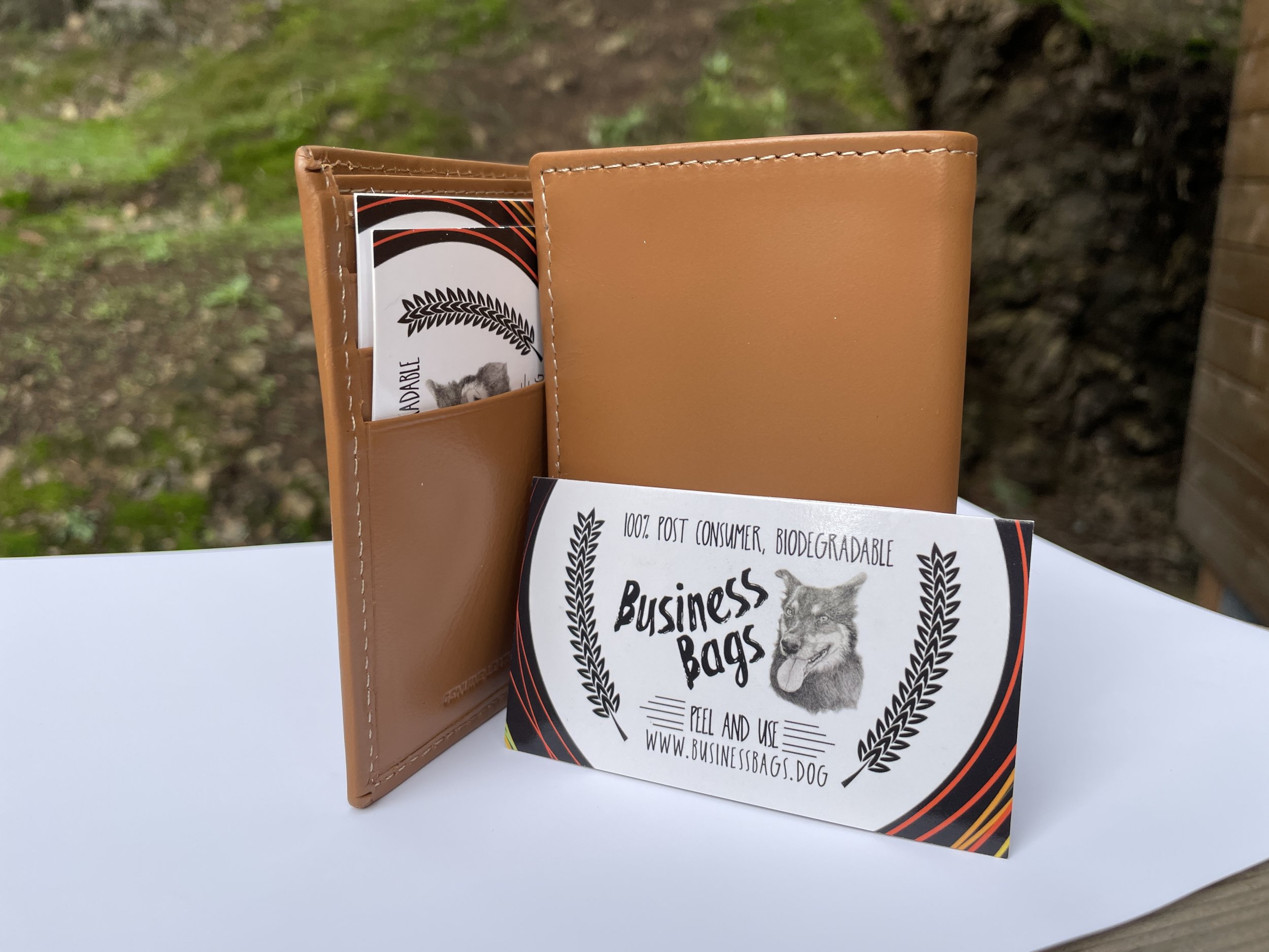 A light brown leather wallet standing open on a white surface outdoors. Next to it is a business card and a rolled-up piece of paper with black and red design, promoting biodegradable, post-consumer business bags with a wolf illustration.