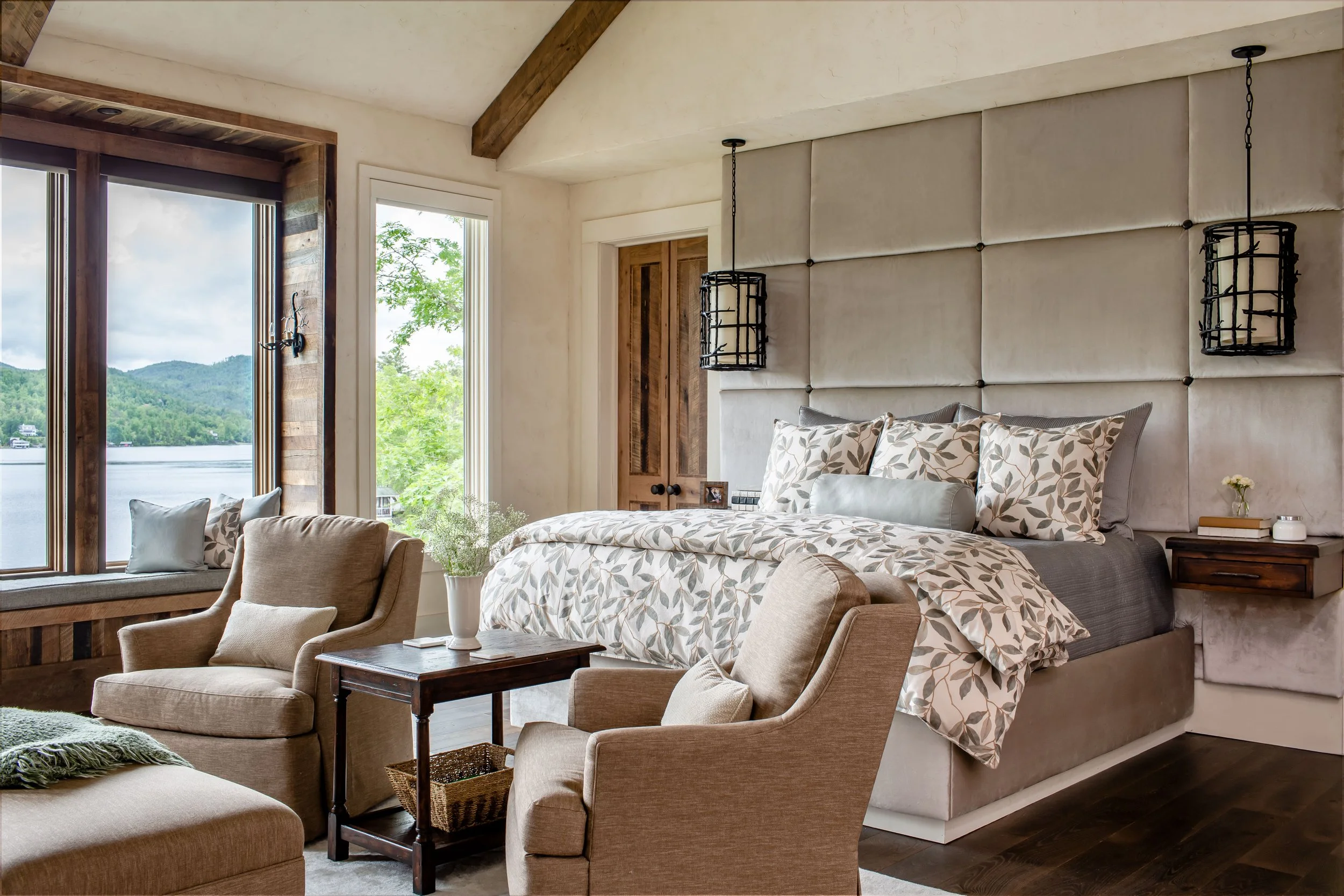 A cozy bedroom with large windows showing a view of a lake and green hills. The room has a bed with a leaf-patterned duvet and pillows, a side table with a small lamp, a beige armchair with a cushion, and hanging black cage-style pendant lights.