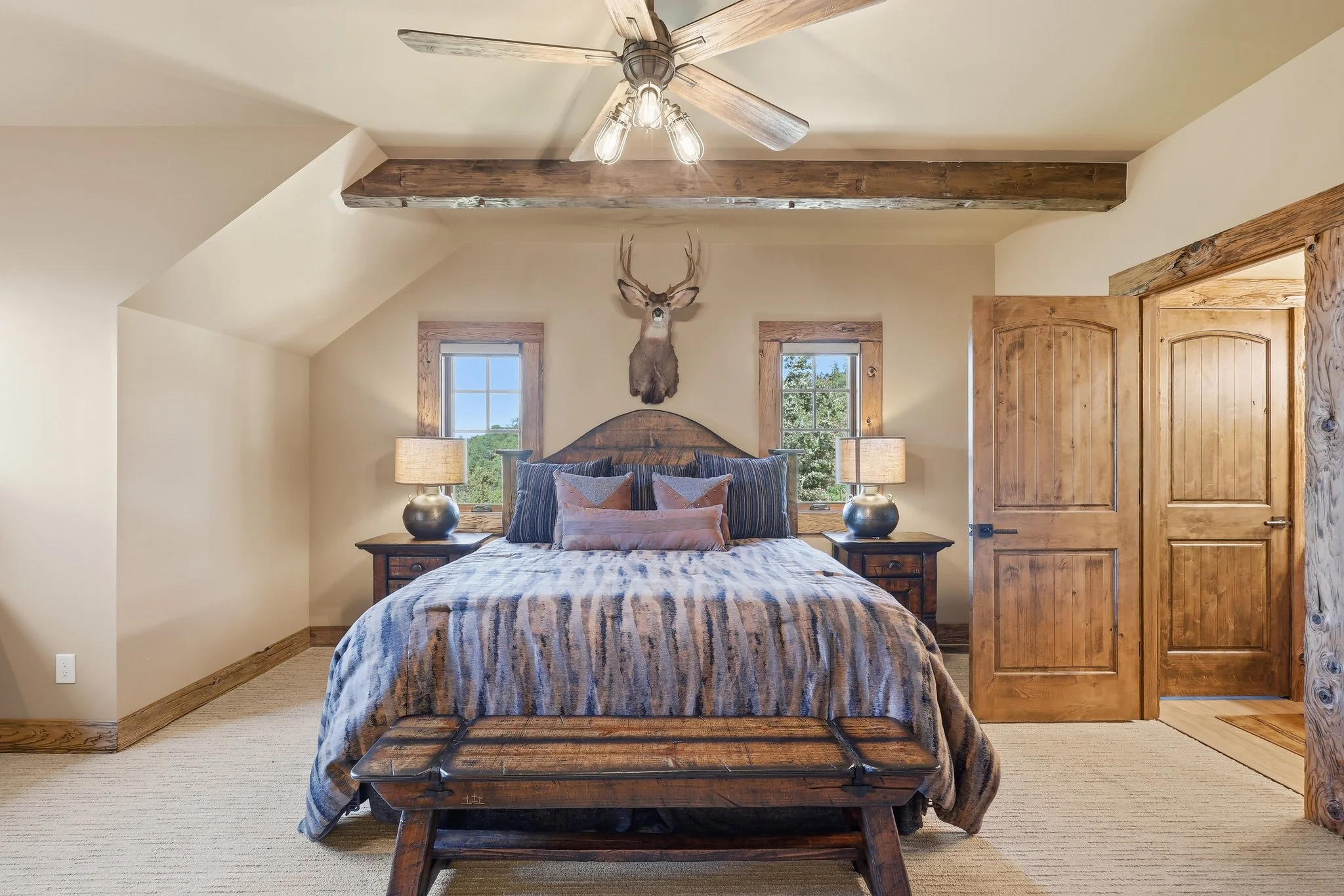A rustic bedroom with a wooden bed, decorative pillows, nightstands with lamps, a mounted deer head, and wooden beams and trim.