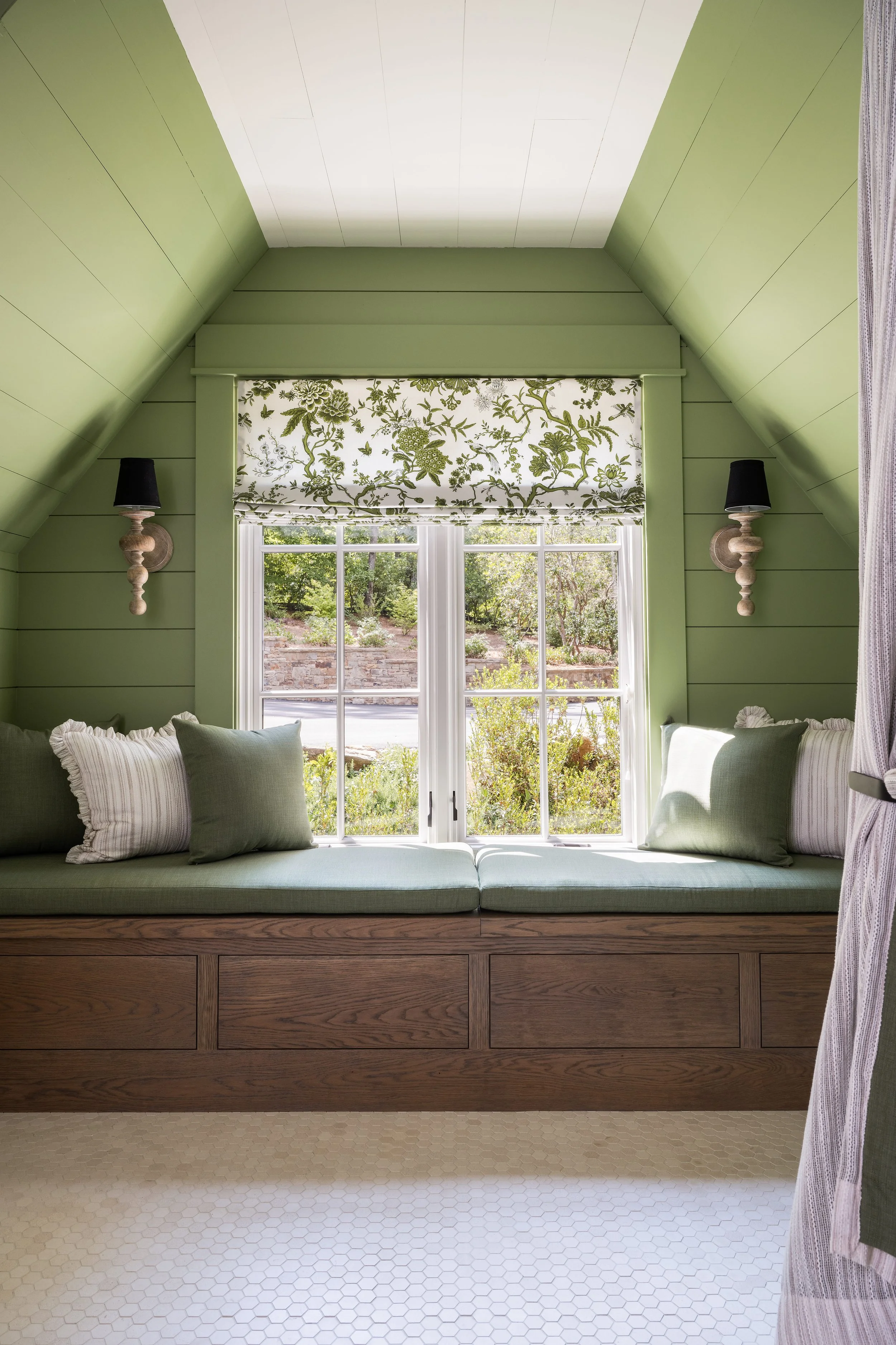 Room with green painted walls and ceiling, featuring a large window with floral patterned Roman shade, two wall-mounted black lamps on carved wooden brackets, and built-in wooden bench with green and beige pillows. Outside the window is a garden with