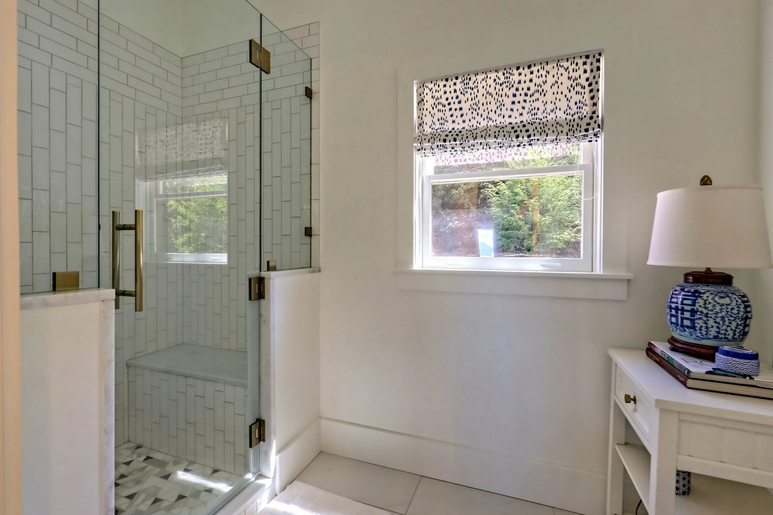 Interior view of a bathroom showcasing a glass-enclosed shower with white subway tile, a window with a patterned Roman shade, and a white side table holding decorative items and books, with a lamp on top.