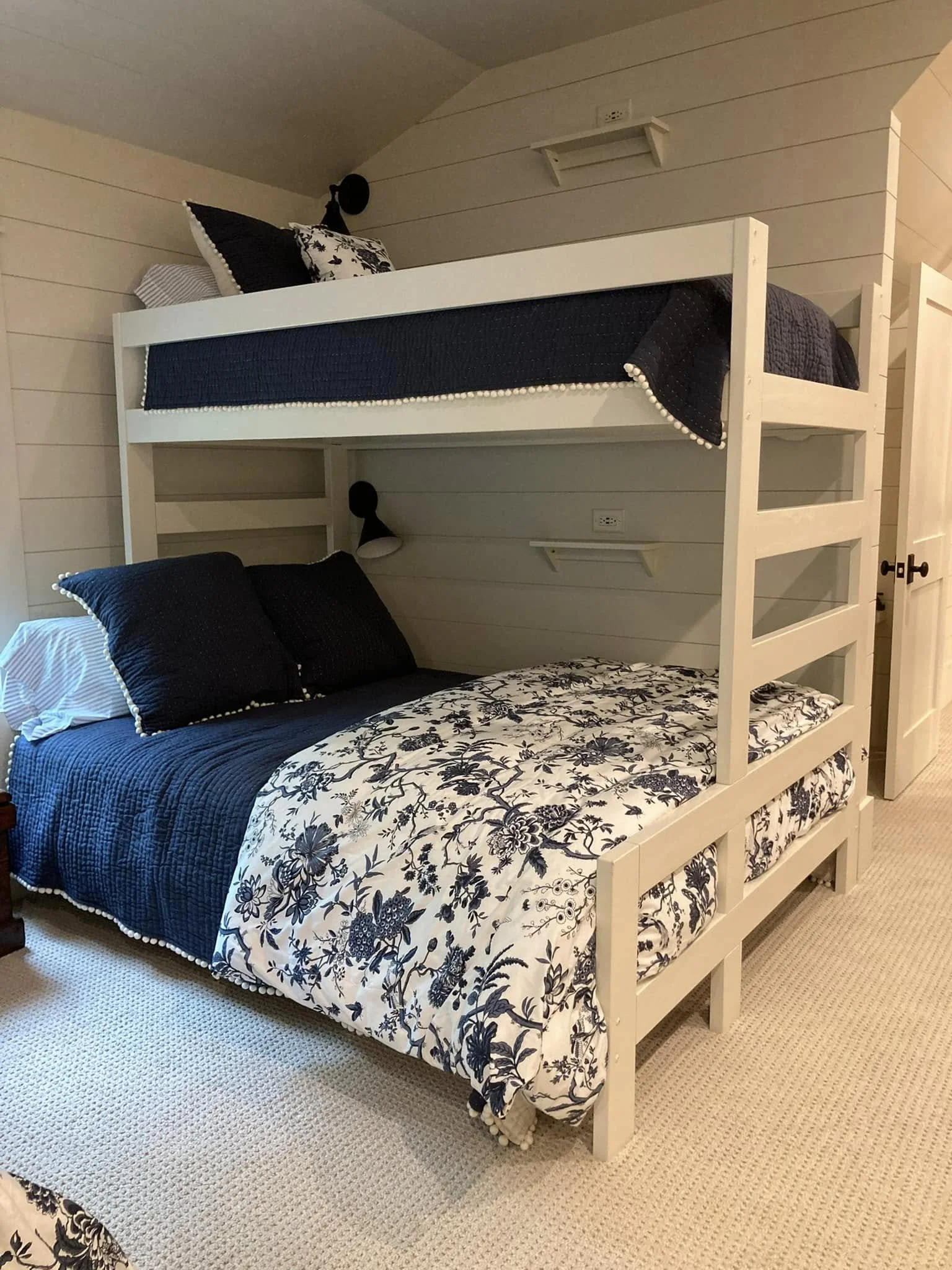 A bedroom with a white bunk bed. The bottom bunk has black pillows and a black and white floral-patterned bedsheet. The top bunk is made with a dark quilt and black pillow, with a smaller decorative pillow. The room has light-colored wooden walls and
