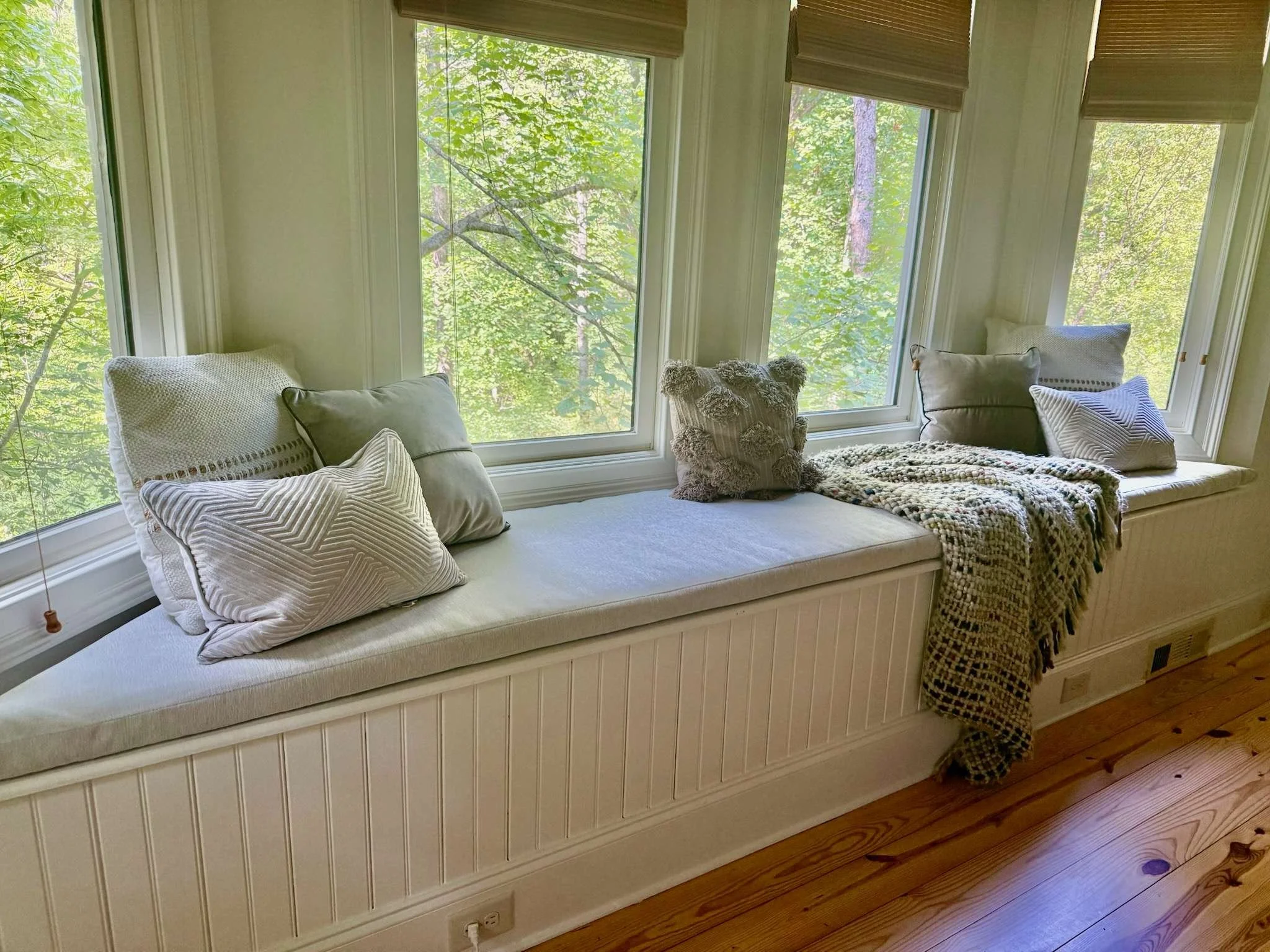 A window seat with several cushions and a soft blanket, overlooking lush green trees outside.