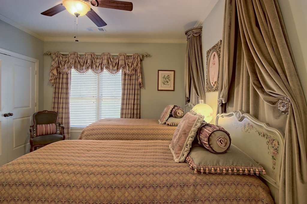 A bedroom with two twin beds, decorative pillows, draped curtains, a ceiling fan, and a chair by the window.