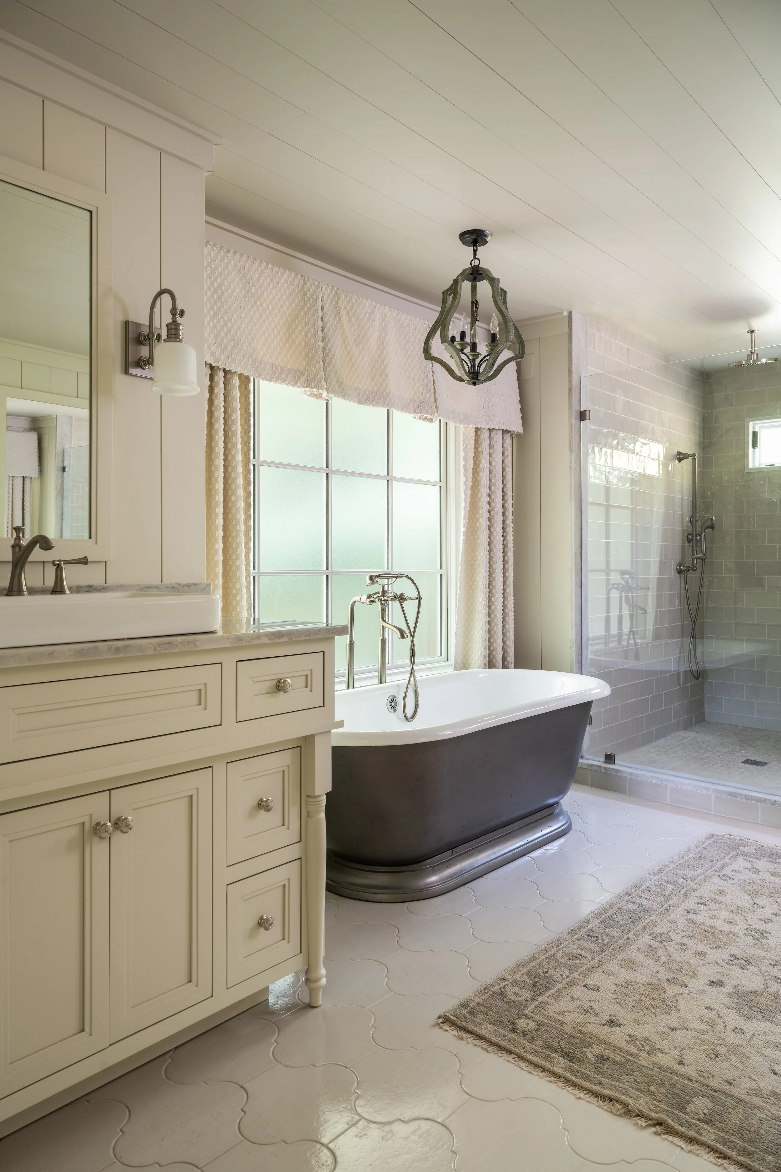 A vintage-style bathroom with a dark gray clawfoot tub near a large window, a white vanity with a marble top and a mirror, a shower area with tiled walls, and a beige patterned rug on white tile flooring.