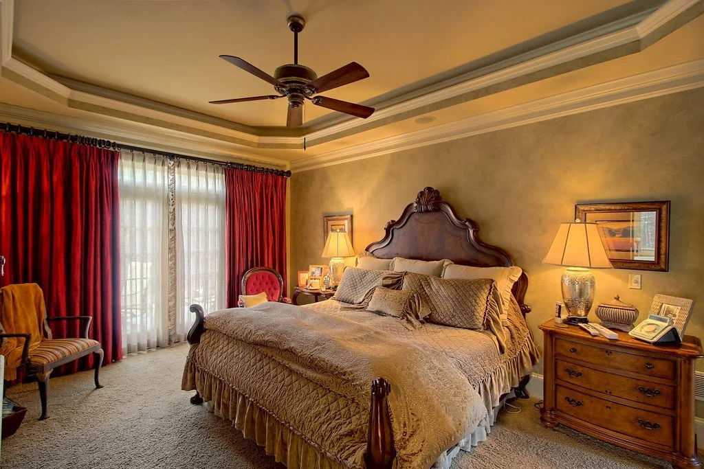 Luxury bedroom with a large bed, ornate wooden headboard, two bedside lamps, and a dresser with a phone and pictures. There are framed pictures on the walls, red curtains covering windows, and a ceiling fan.