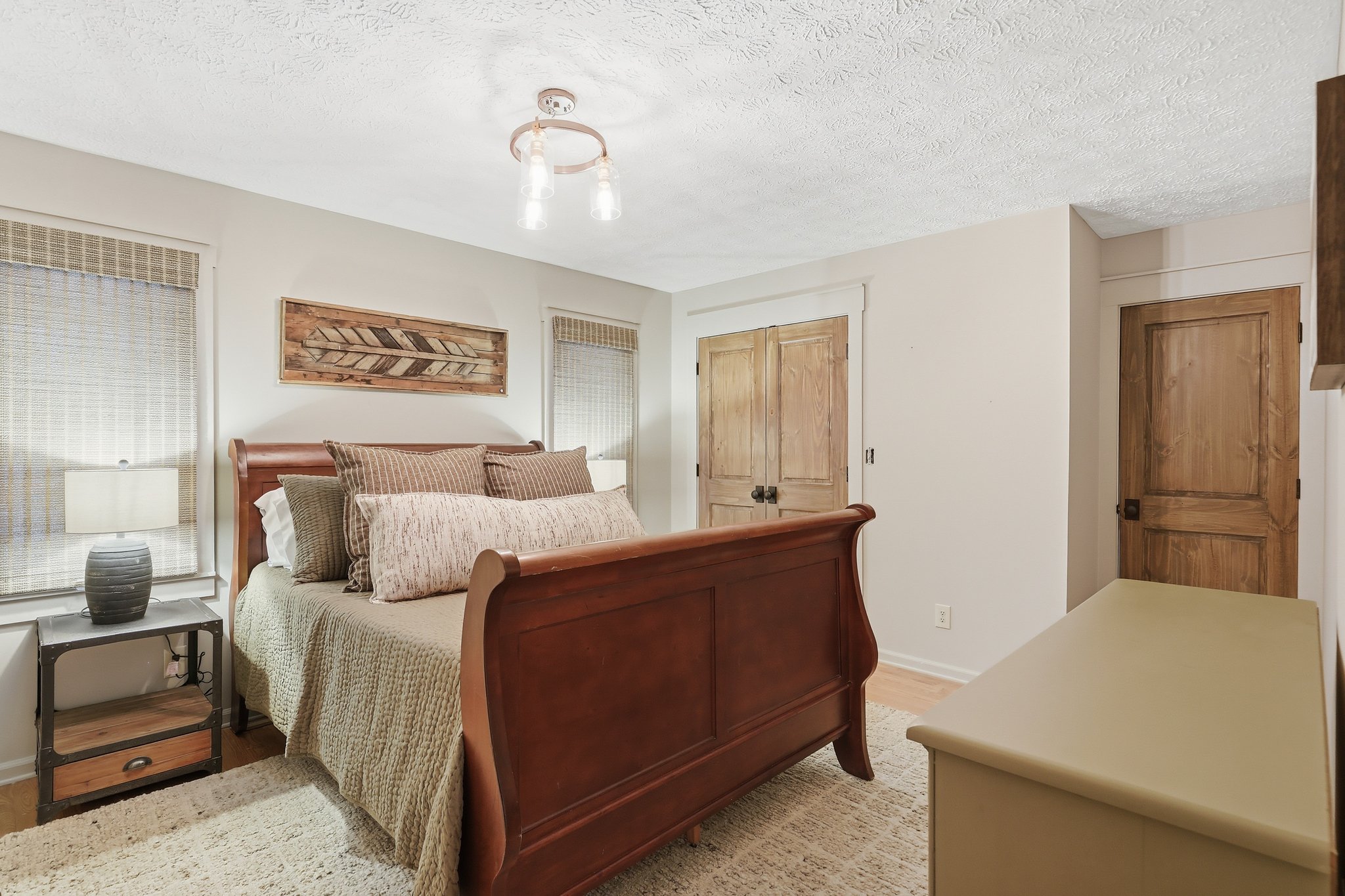 A bedroom with a wooden bed frame, beige bedding, multiple pillows, a nightstand with a lamp and vase, two windows with blinds, wooden closet doors, a white textured ceiling, and a ceiling light fixture.