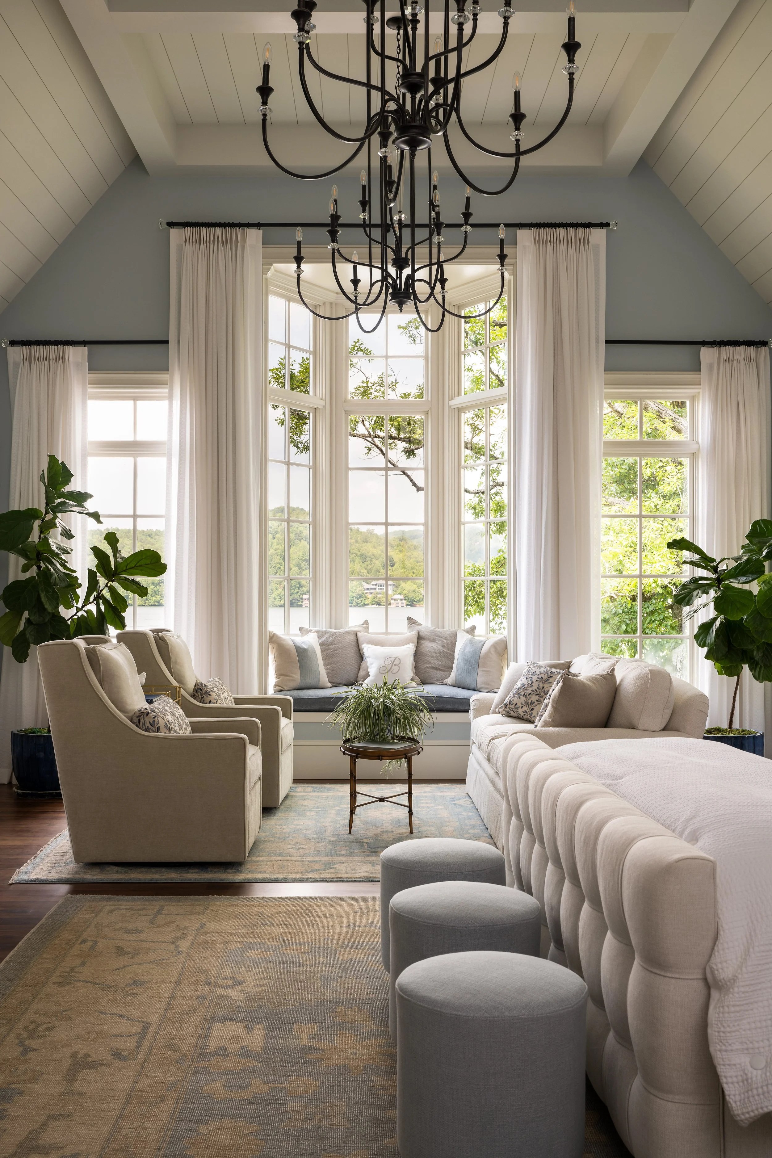 A spacious living room with a large bay window, surrounded by white curtains, and a chandelier hanging from the ceiling. The room has light-colored furniture, including a sofa, armchairs, and small ottomans, as well as potted plants and a view of tre