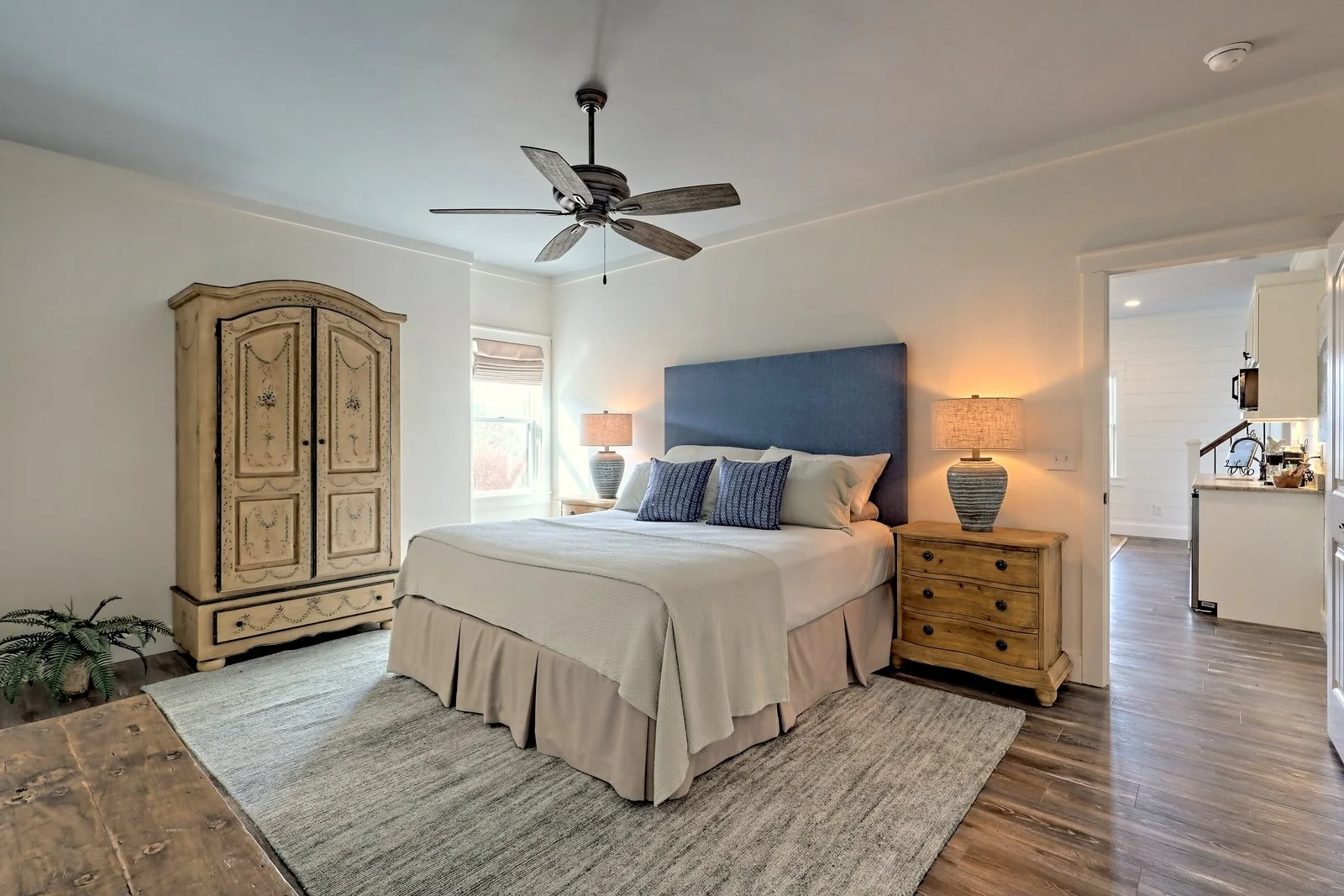 A bedroom with a large bed featuring a blue headboard, beige bedding, and blue pillows. There are two wooden nightstands with lamps on each side of the bed, and a wooden armoire on the left. The room has a ceiling fan, a window with a blind, and a be