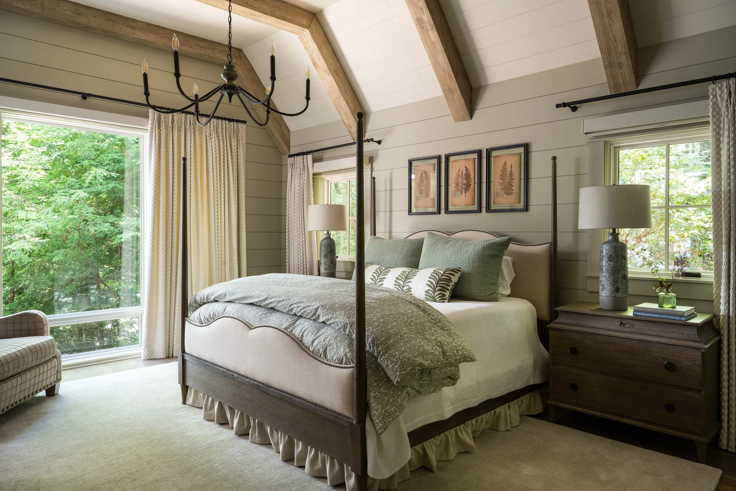 A bedroom with a four-poster bed, a nightstand with lamps and books, framed botanical art on the wall, large windows with curtains, and a ceiling chandelier, surrounded by green foliage outside.