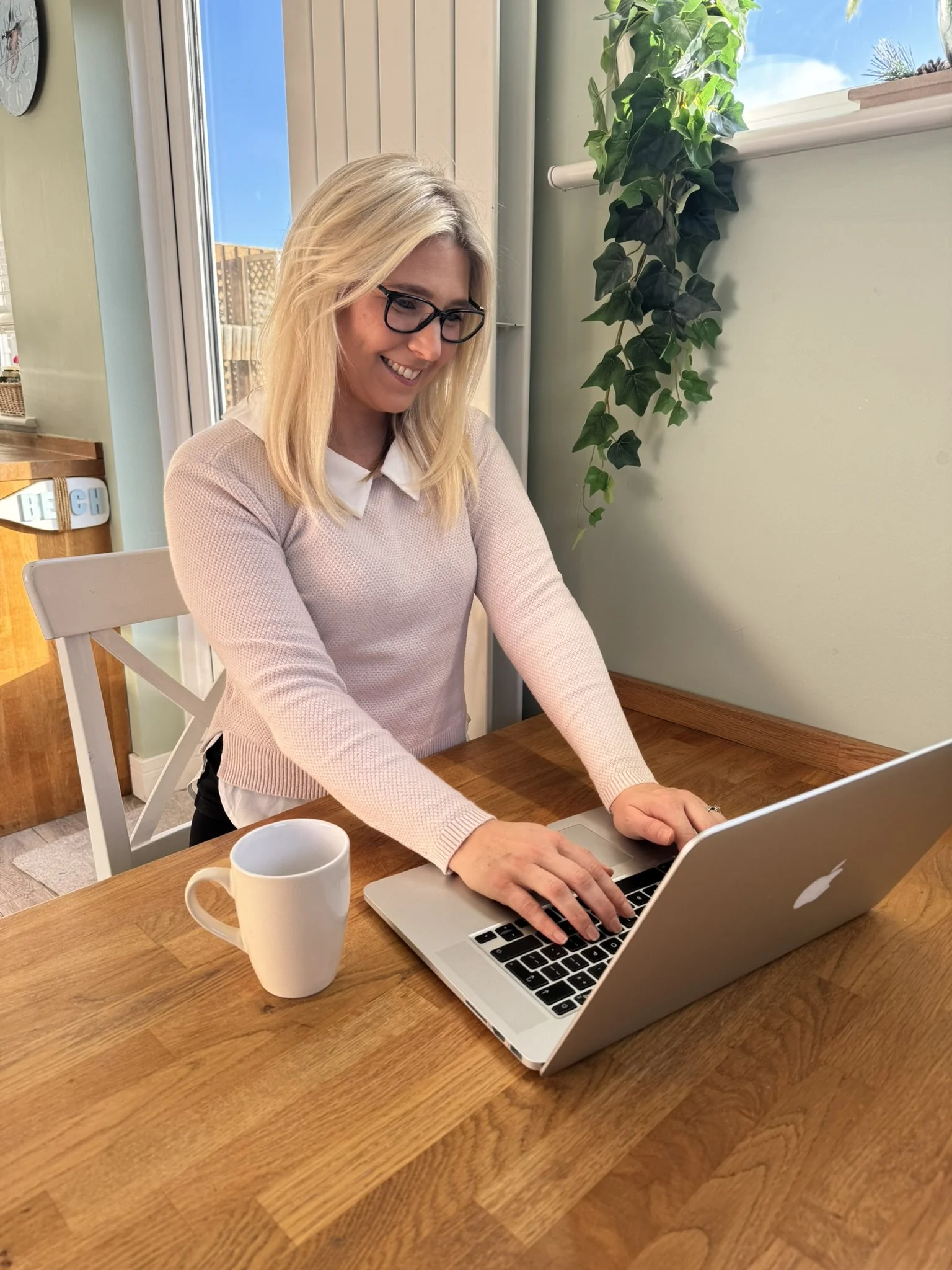 A woman with blonde hair, glasses, and a light pink sweater working on a silver laptop at a wooden table, with a white mug nearby, in a bright room with a large window and green plants.