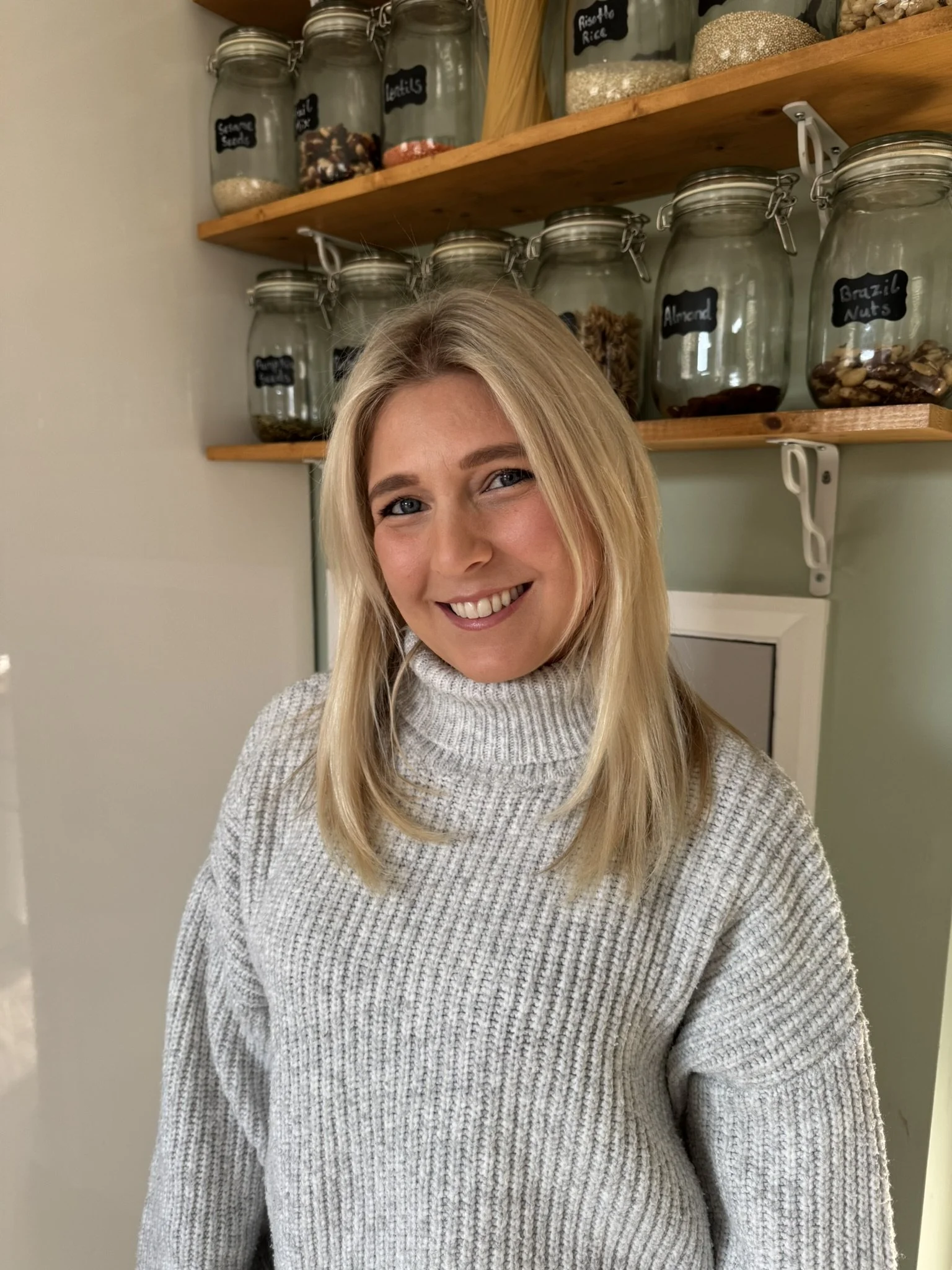 A woman with blonde hair wearing a warm turtleneck sweater, smiling in front of a shelf filled with glass jars containing various whole food goods.