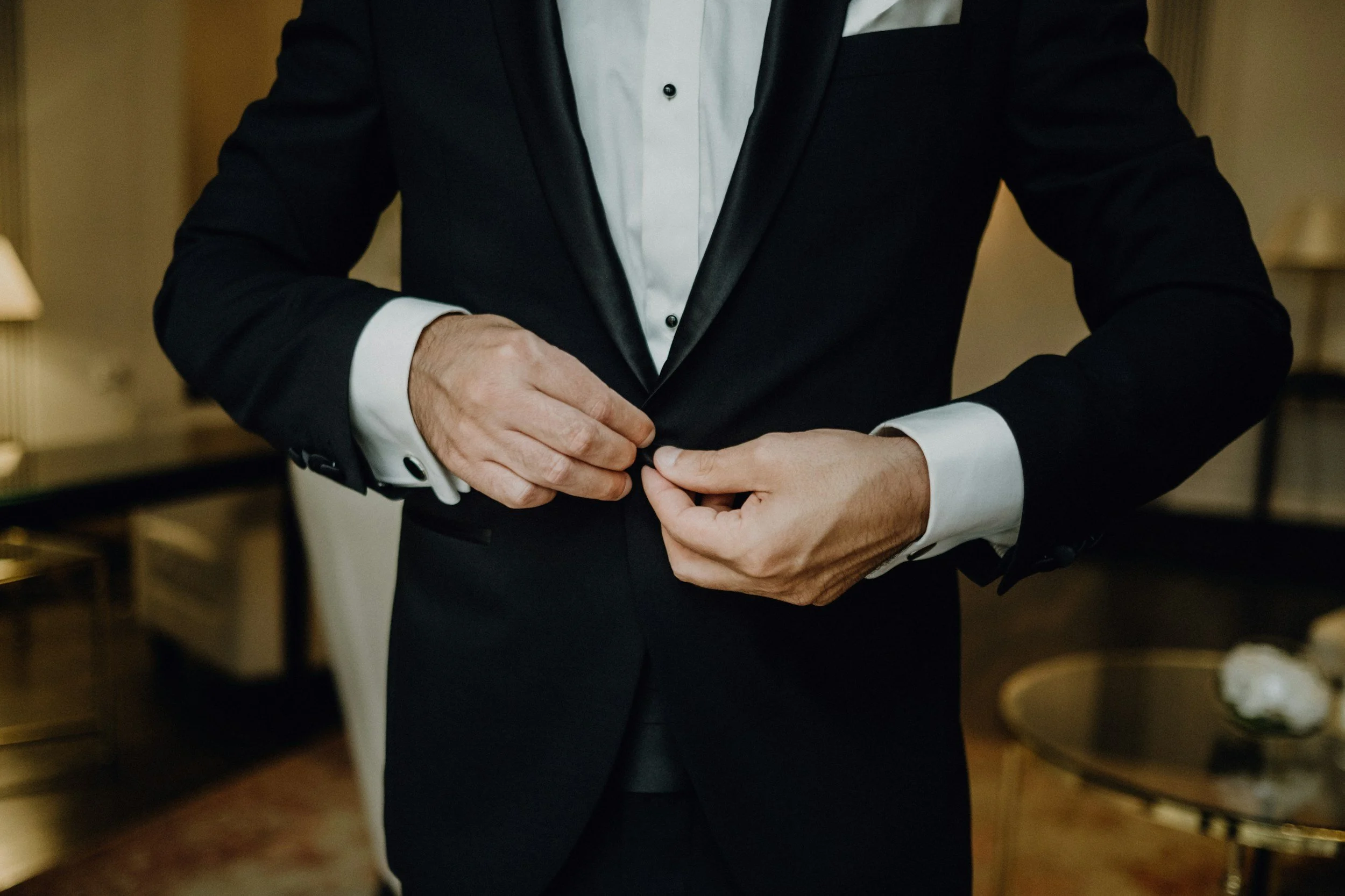 A man in a tuxedo is buttoning the front of his jacket in a warmly lit room.