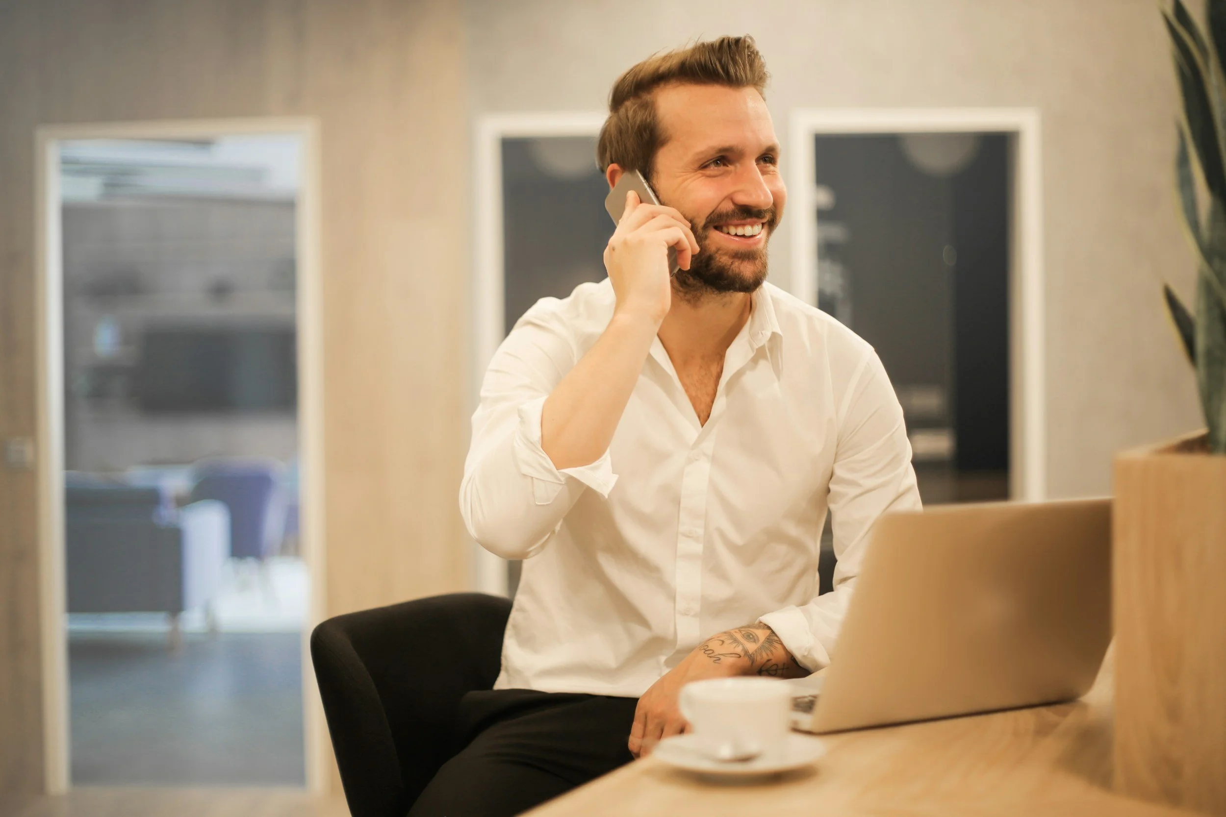 A man with a beard and tattoos, wearing a white shirt, smiling while talking on a mobile phone at a desk with a laptop, coffee, and a plant in an office setting.