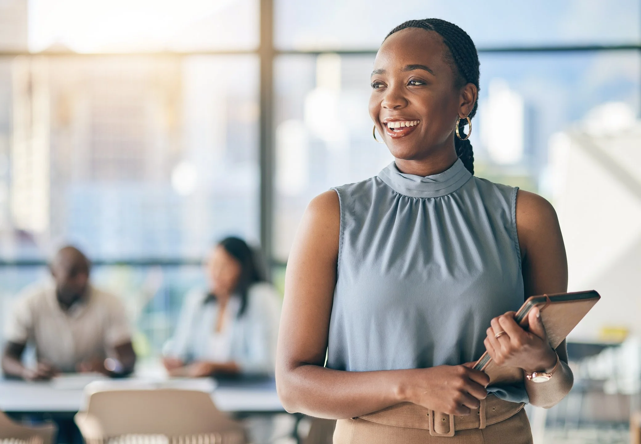 A smiling Black woman in a sleeveless gray blouse holding a clipboard, in a modern office with large windows and blurred colleagues in the background.