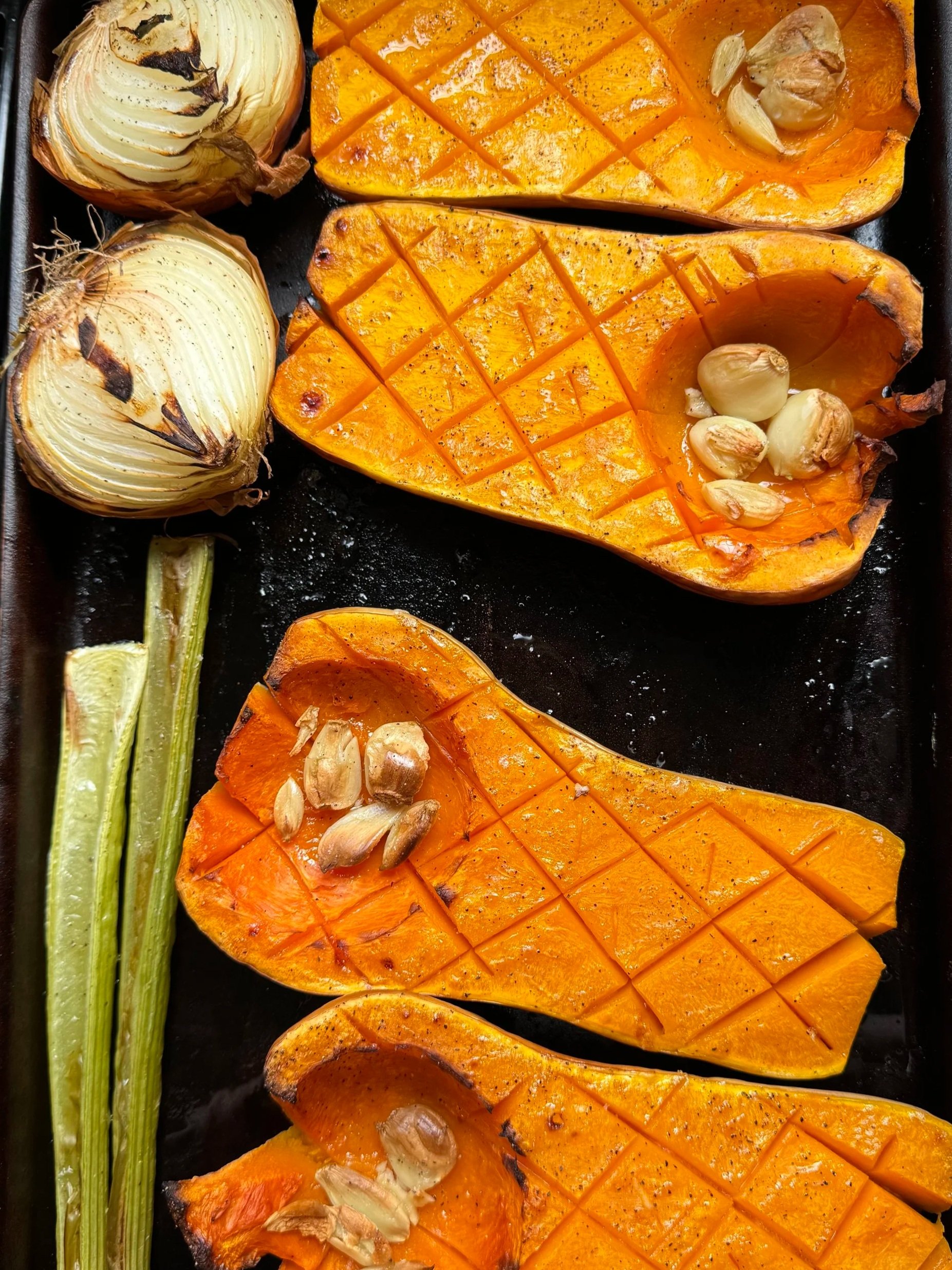 Halved butternut squash slices with seeds, two roasted onions, and celery stalks on a black baking tray.