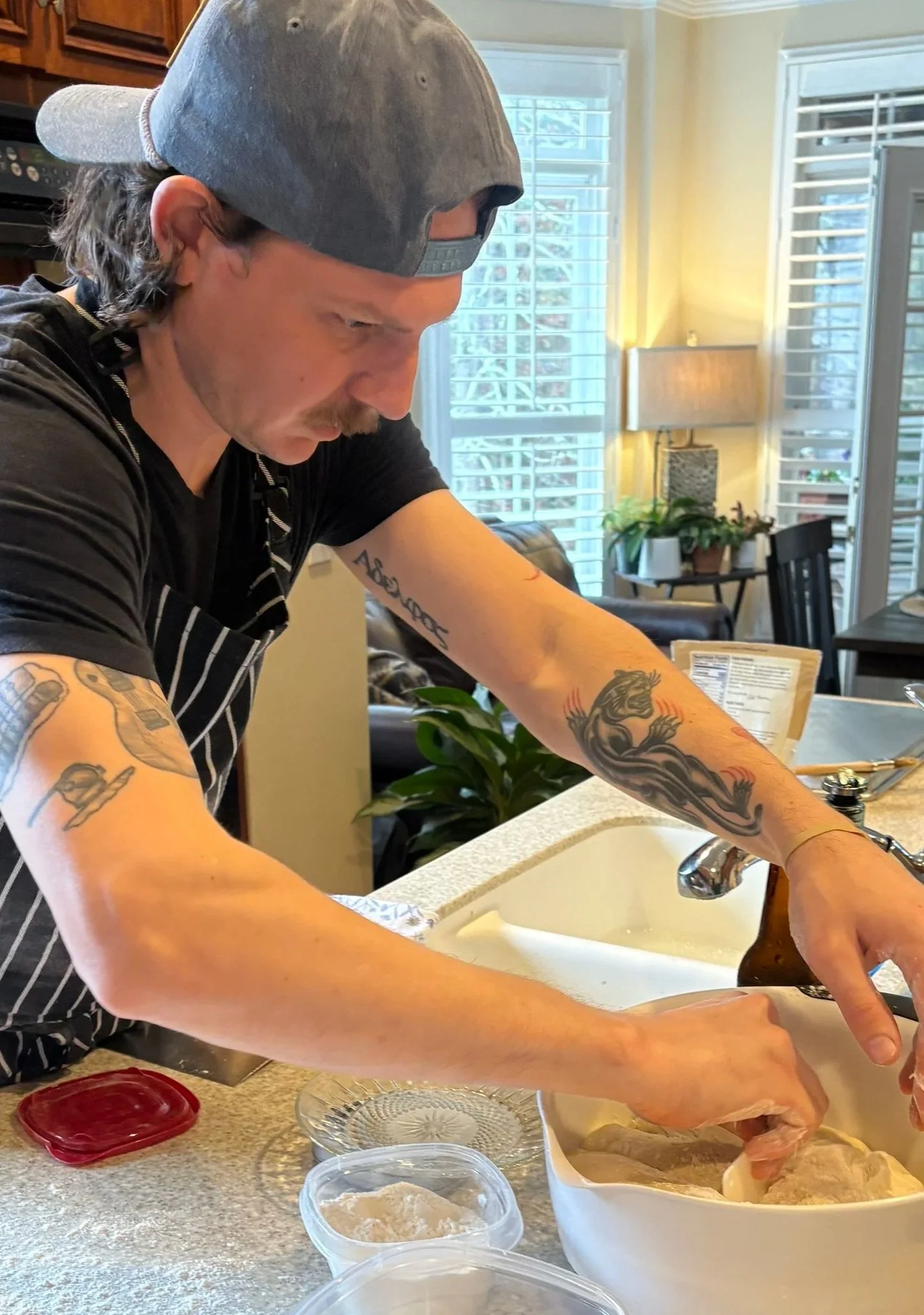 A man with tattoos on his arms and wearing a gray baseball cap and a black t-shirt, standing at a kitchen sink preparing chicken. The kitchen has large windows with white blinds, a lamp with a beige shade, a small table with potted plants, and dining chairs in the background.