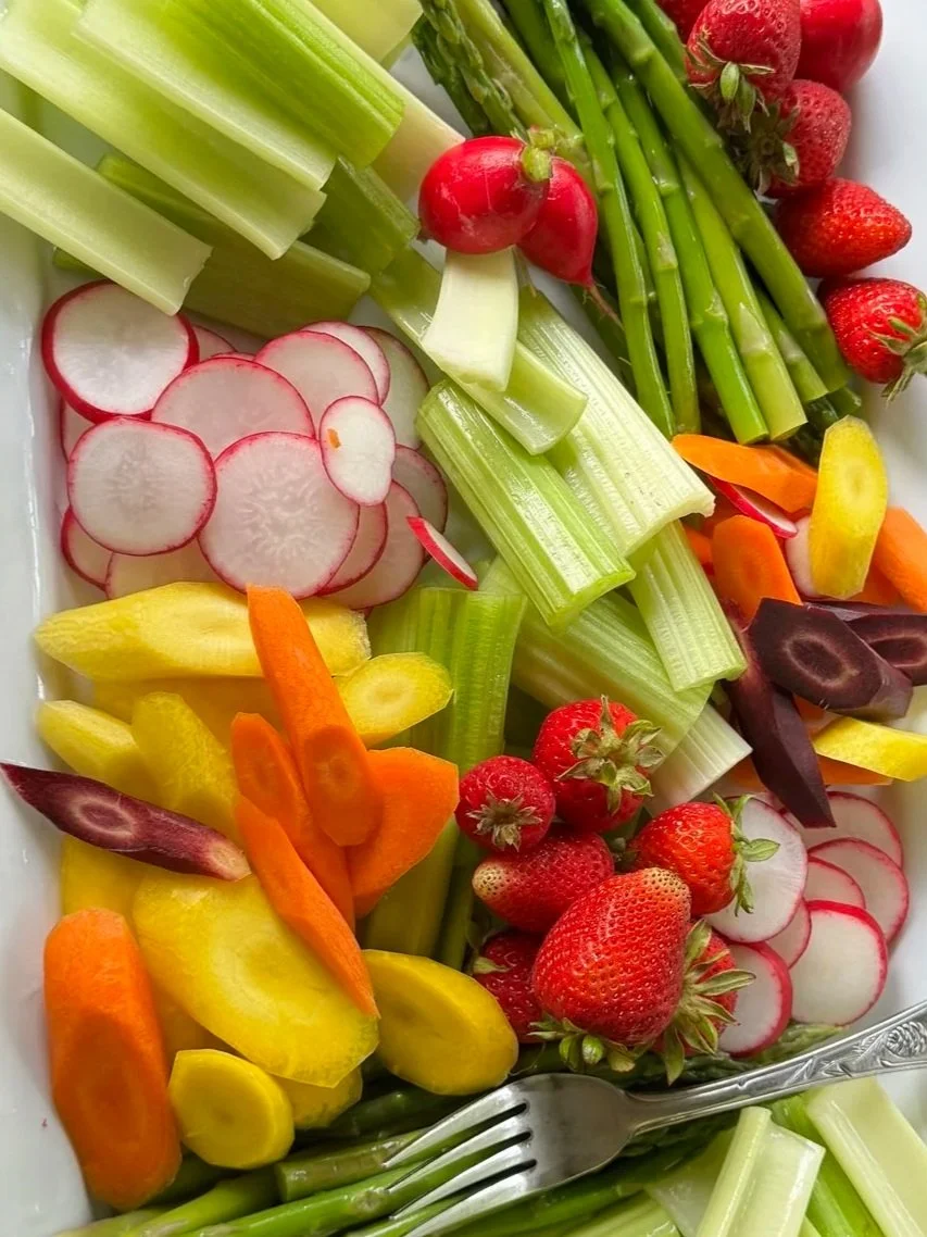 Fresh sliced radishes, strawberries, celery, carrots, yellow bell peppers, and green asparagus on a white platter.