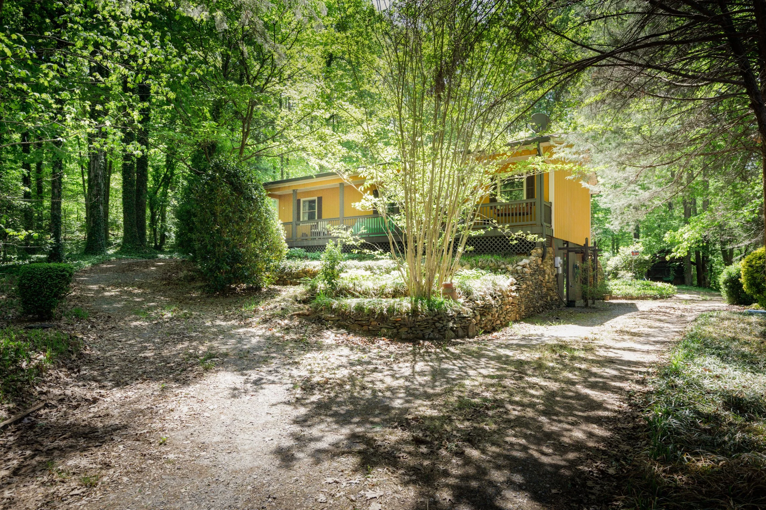A yellow house with a porch, surrounded by tall green trees with sunlight filtering through the leaves, and a dirt path leading toward the house.