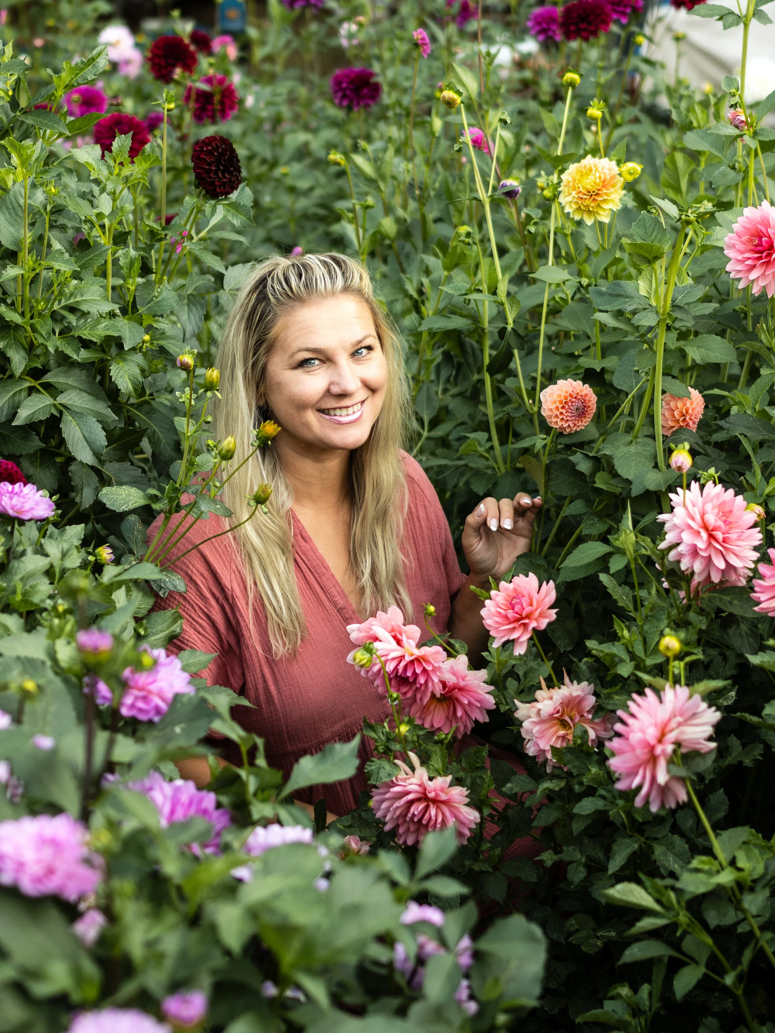 A woman with long blonde hair smiling among pink and purple dahlias in a garden.