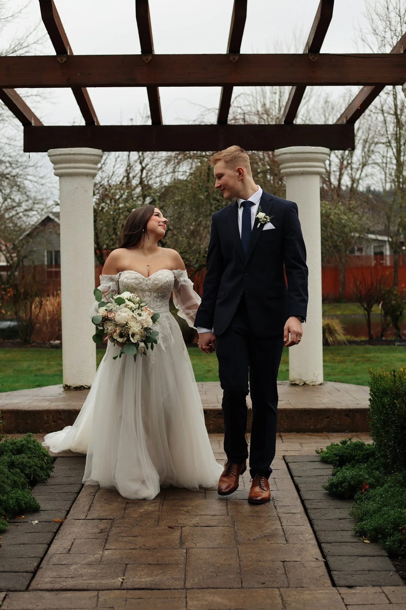 A bride and groom holding hands and looking at each other, standing outdoors under a wooden pergola with white columns on a cloudy day.