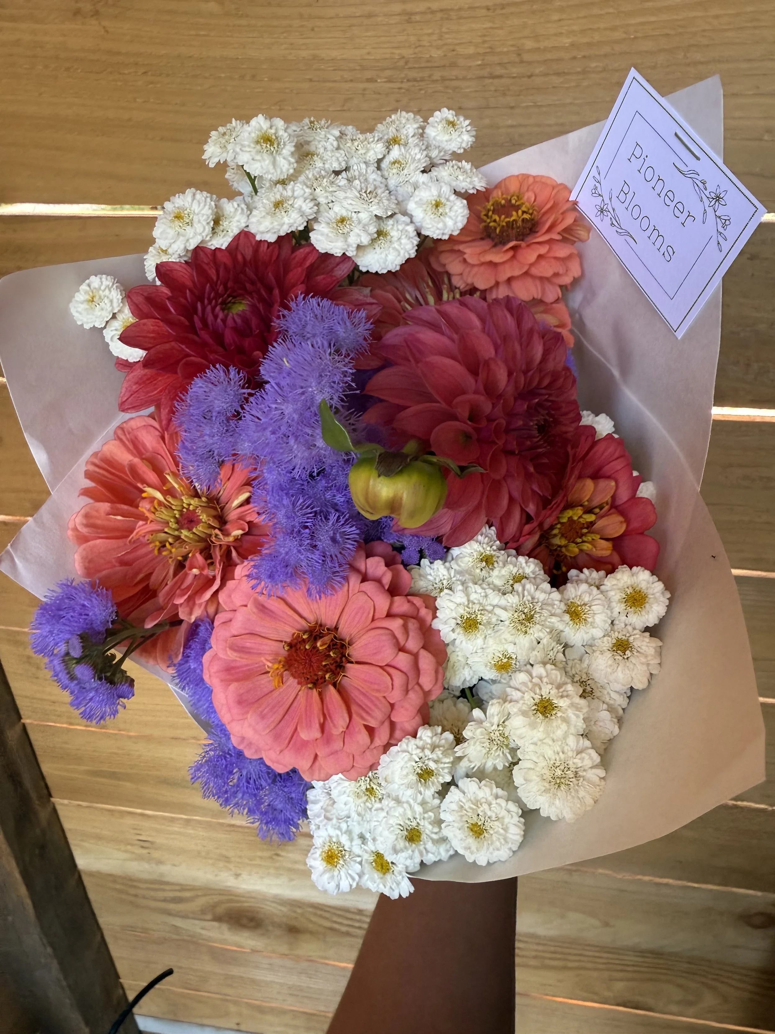 A colorful bouquet of flowers with white, pink, red, and purple blooms, wrapped in white paper, with a small card that reads 'Pioneer Blooms'.