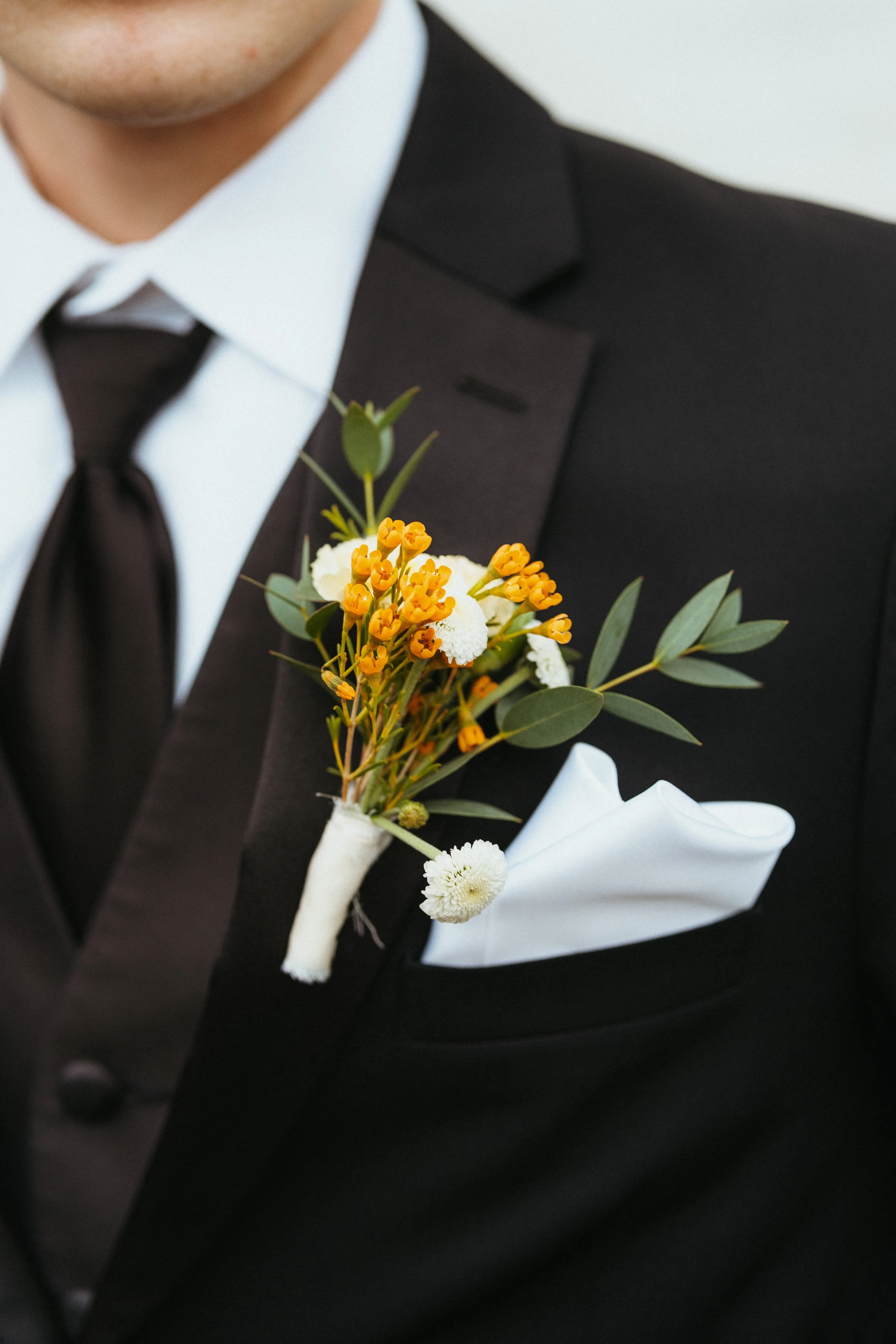 Close-up of a man's shoulder in a black tuxedo with a white dress shirt, black tie, and a white pocket square, with a boutonniere made of yellow and white flowers and green leaves pinned to the lapel.