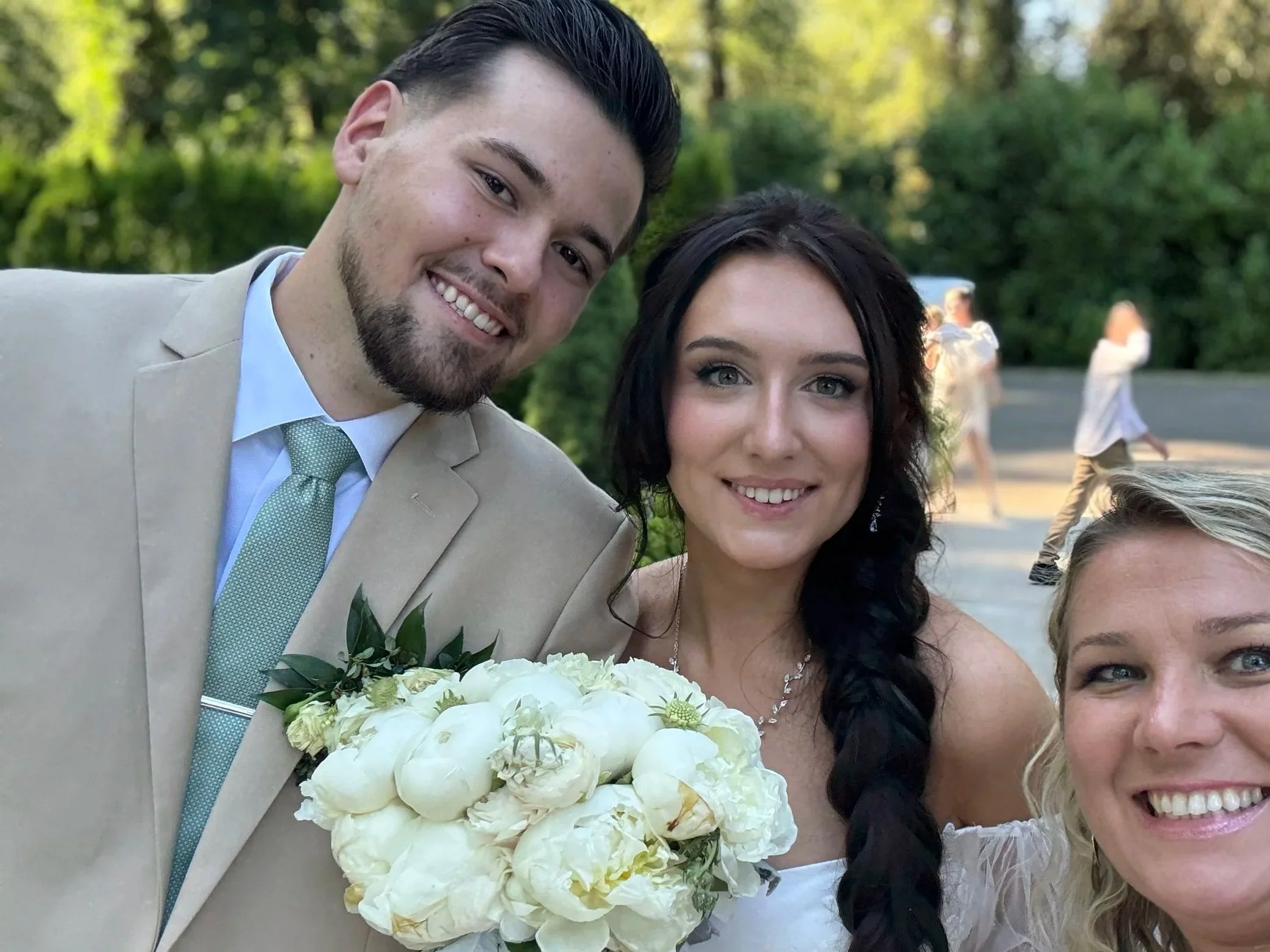 Three people, two women and one man, smiling for a photo outdoors. One woman is holding a bouquet of white flowers. The man is dressed in a beige suit with a light green tie, and the women are dressed in white. There are trees and other people in the