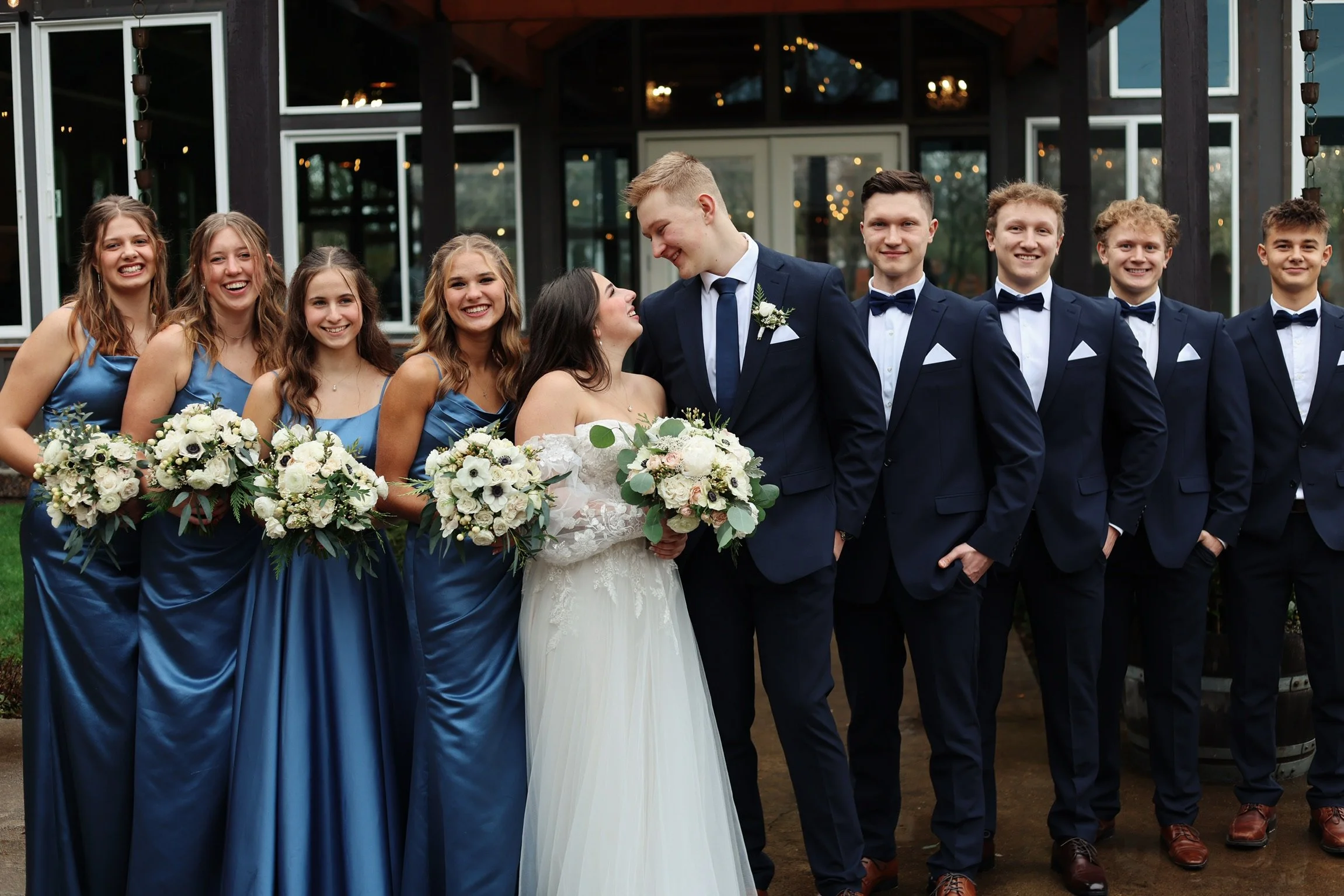 A wedding party with eight people standing outdoors in front of a building with large windows. The bride and groom are in the center, smiling and looking at each other, holding bouquets. The bride is wearing a white dress, and the groom is in a navy 